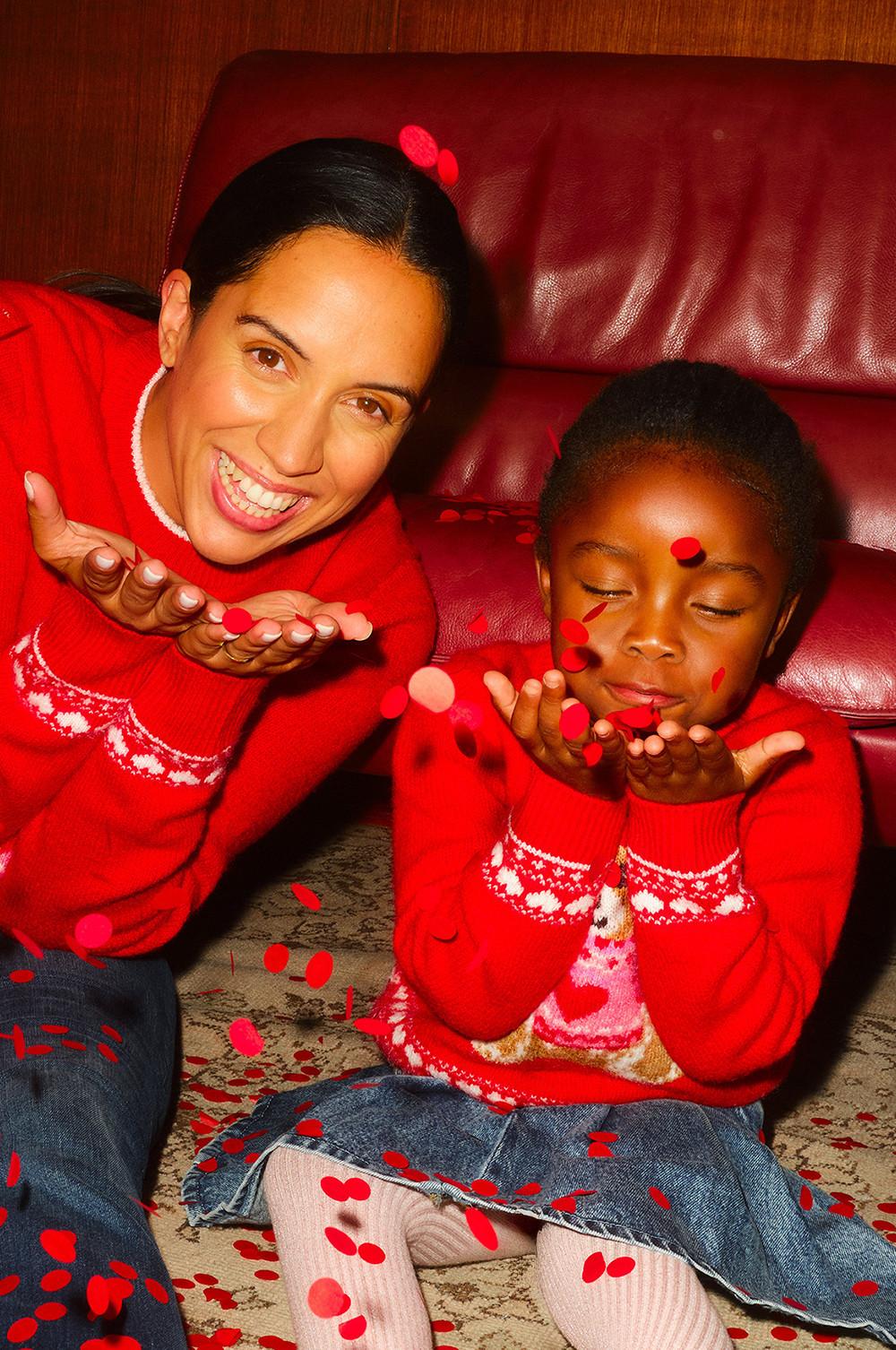 Two people wearing matching red Christmas jumpers with festive patterns, blowing red confetti from their hands.