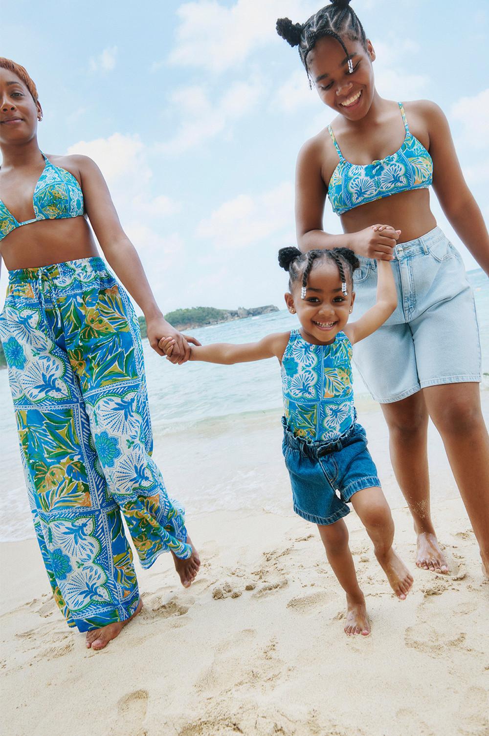 Una adulta y dos niñas con prendas de baño estampadas en azul y amarillo. La adulta con bikini combinado con pantalones estampados a juego, una de las niñas con bikini combinado con vaqueros cortos y la otra niña con un traje de baño azul estampado y vaqueros cortos azul oscuro.