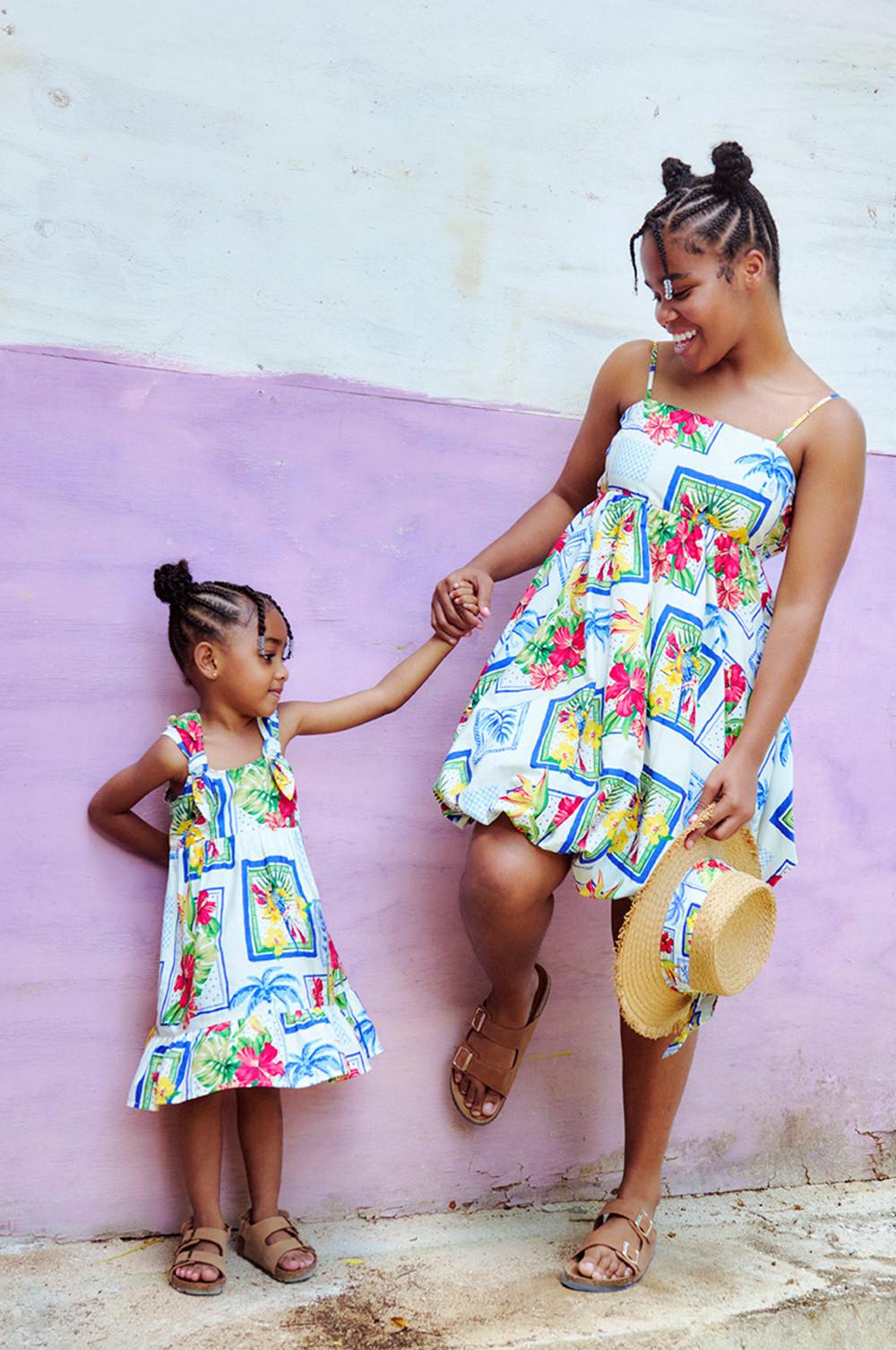 Dos modelos infantiles con vestidos con estampado de postales tropicales a juego, sandalias en color tostado y sombrero de paja.