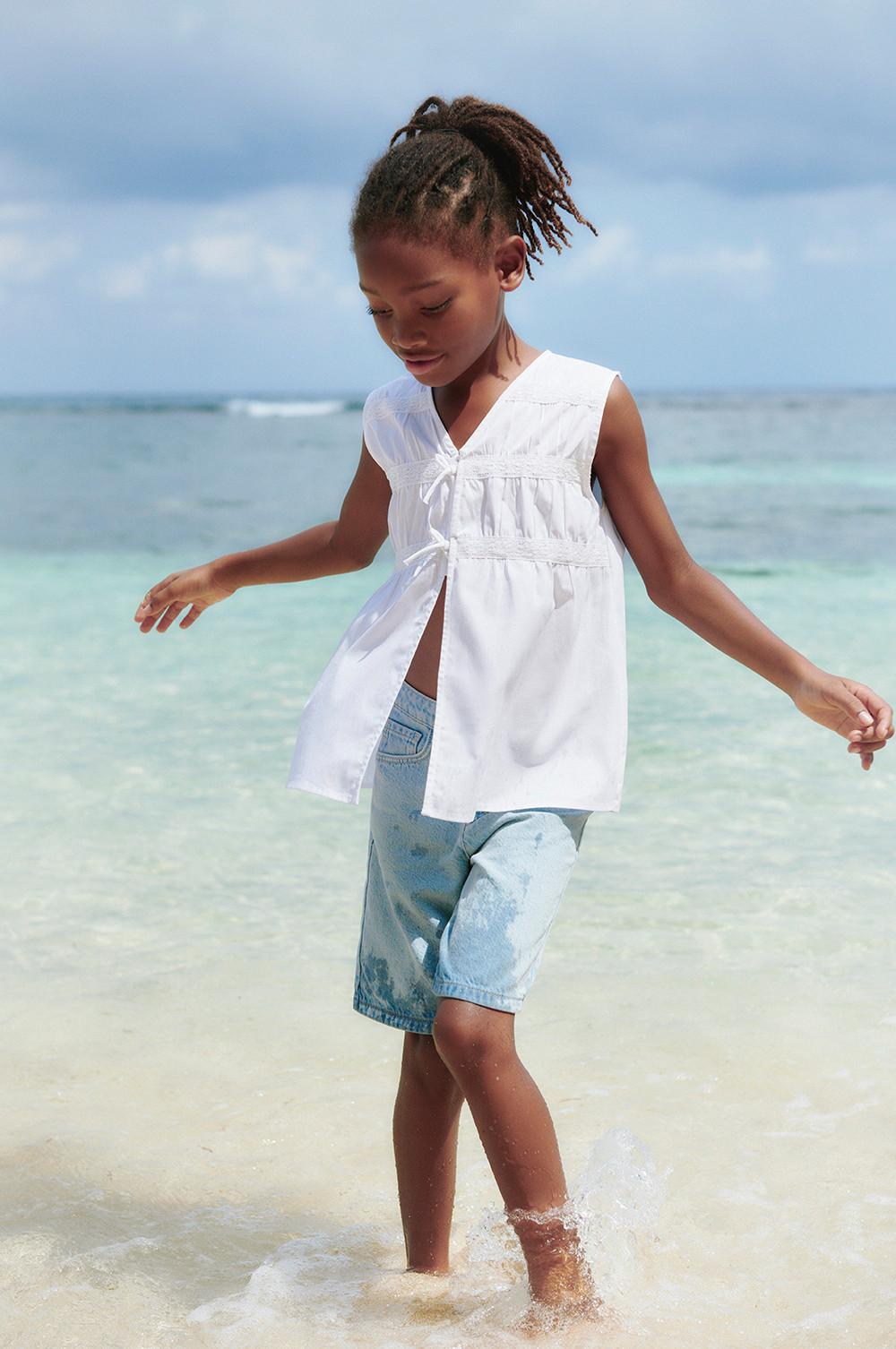 Modelo infantil con blusa blanca sin mangas con lazada delantera y vaqueros cortos en azul claro.
