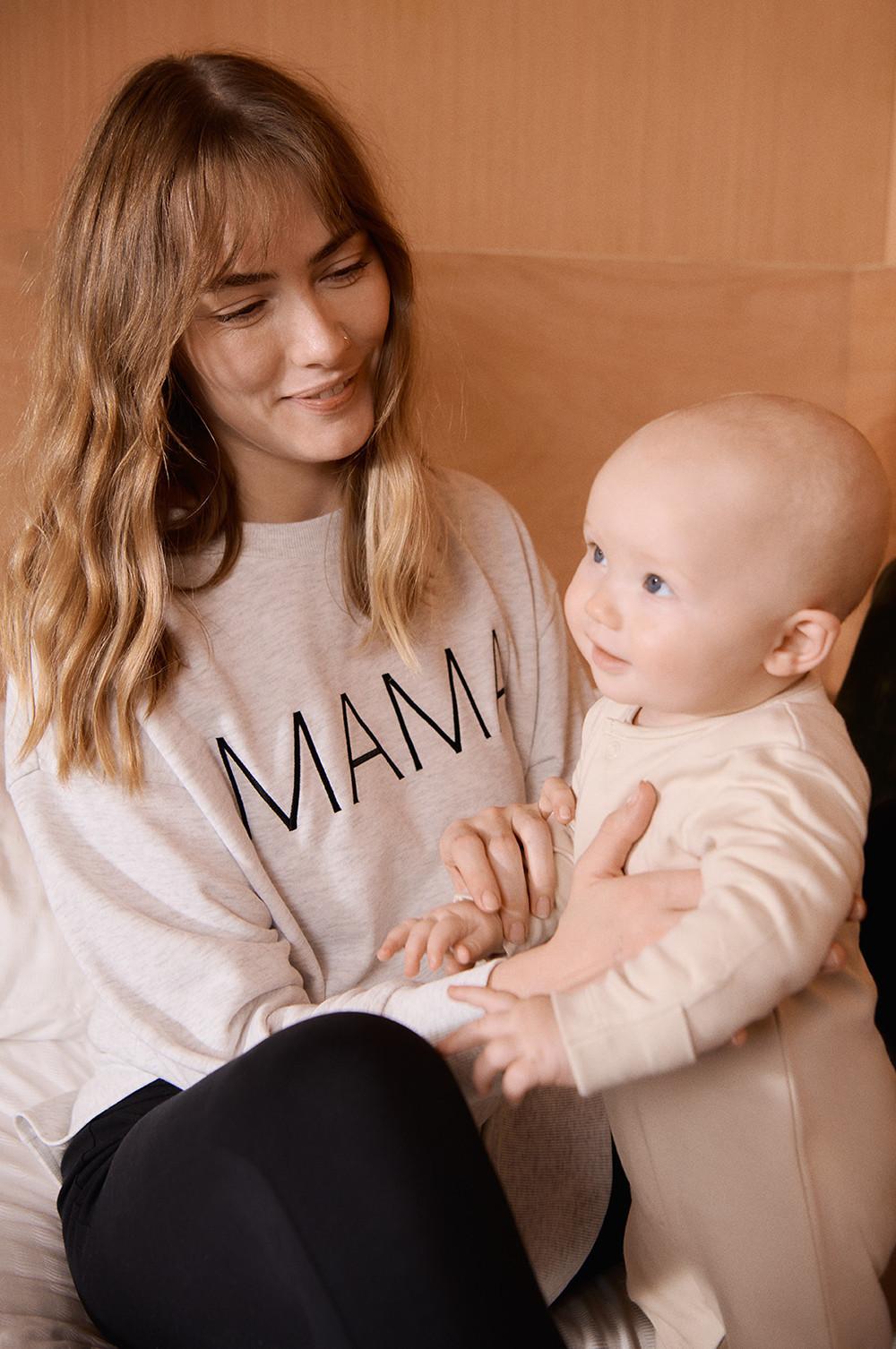 Mannequin portant un sweat-shirt crème orné de l'inscription « MAMA » brodée en noir sur le devant, associé à un legging noir, et tenant un bébé vêtu d'une grenouillère blanche.