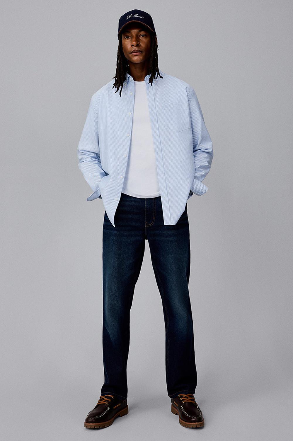 Personne portant une chemise bleu clair à rayures sur un t-shirt blanc et un jean droit foncé avec des chaussures bateau marron.