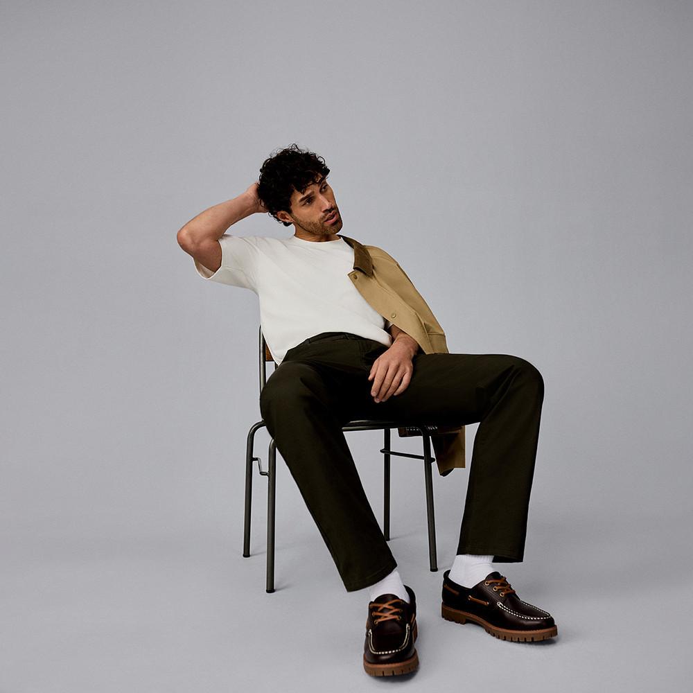 Personne assise sur une chaise portant un t-shirt, une veste beige, un pantalon foncé et des chaussures bateau marron.