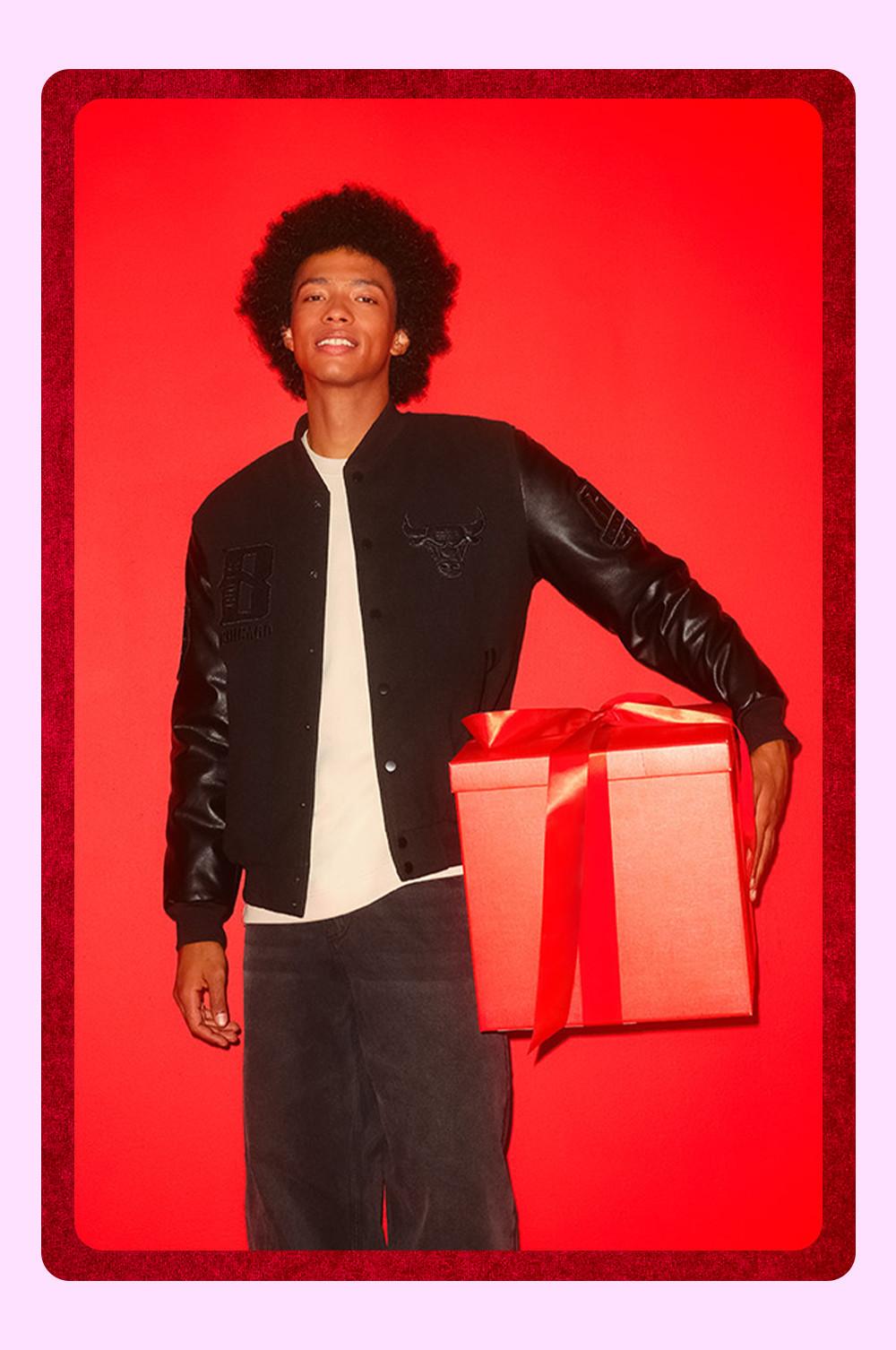 Persona che indossa una giacca in stile college nera e jeans che tiene in mano un grande pacco regalo rosso con un nastro su sfondo rosso.