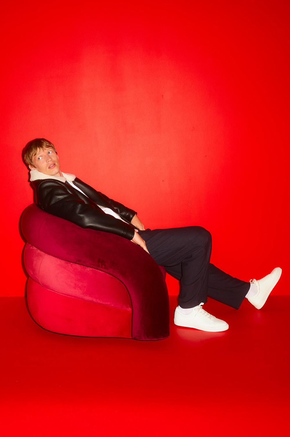 Persona con giacca di pelle nera seduta su una sedia rivestita in velluto rosso su uno sfondo rosso brillante.