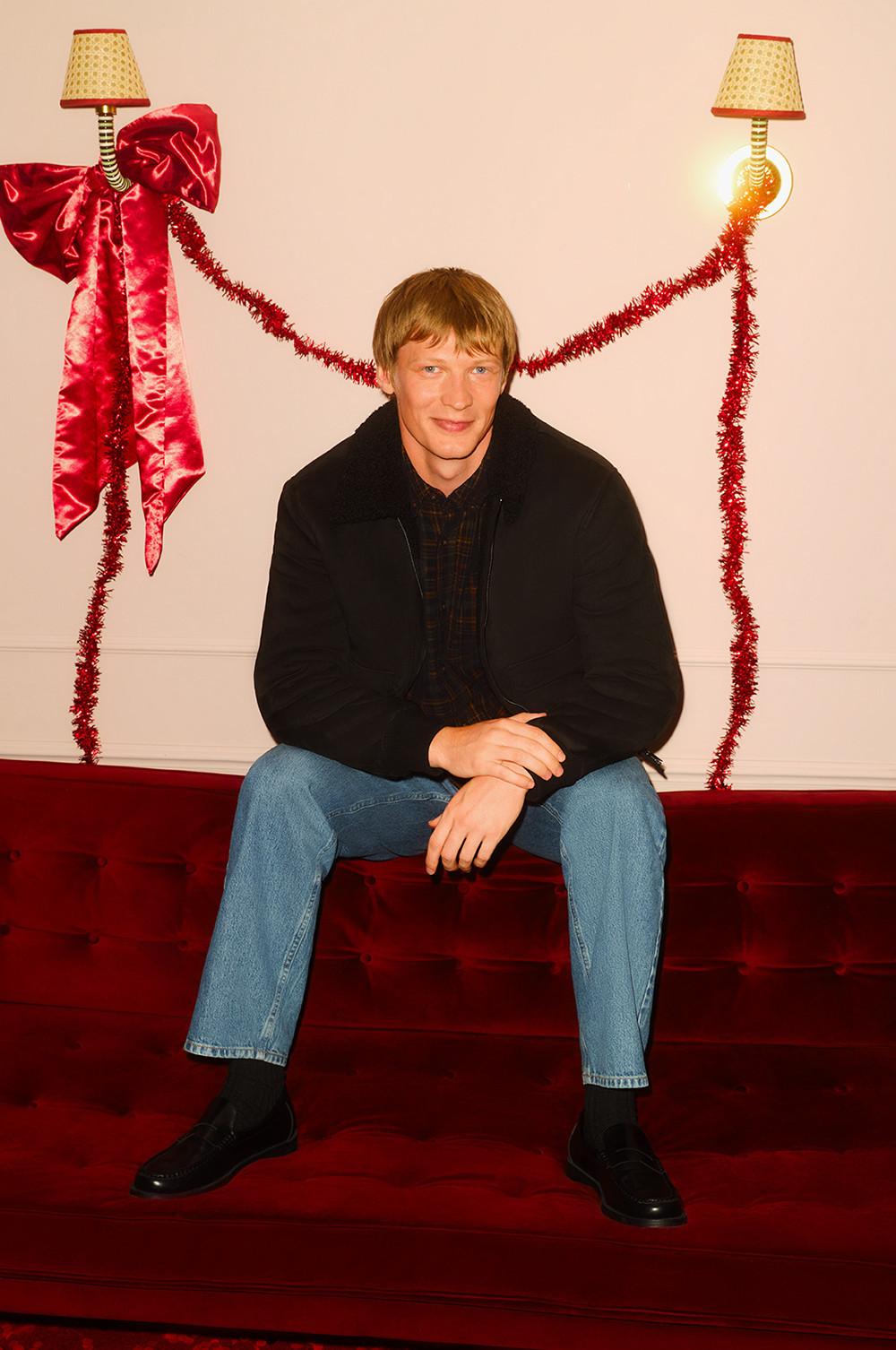 Persona seduta su un divano rosso che indossa una giacca nera e jeans, con decorazioni festive e lampade intorno.