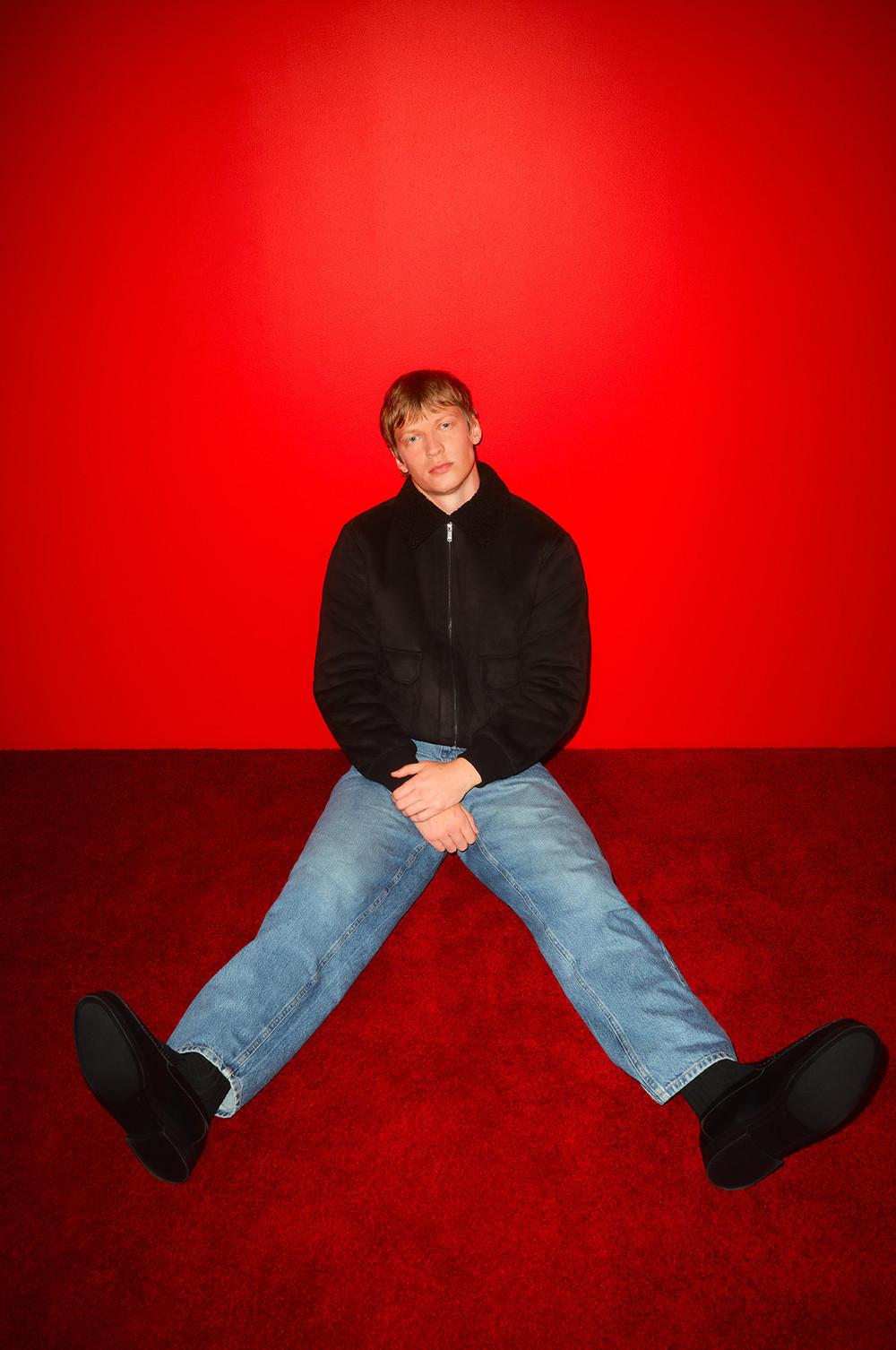 Persona che indossa una giacca nera con zip e jeans blu seduta con le gambe divaricate su uno sfondo rosso brillante.