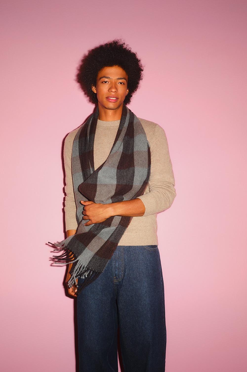 Persona che indossa un maglione beige con sciarpa a quadri blu e grigi e jeans su sfondo rosa.