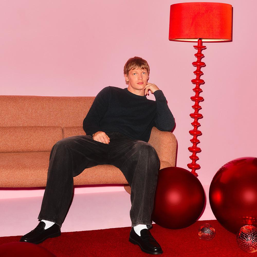 Persona con outfit nero seduta su un divano marrone con una caratteristica lampada da terra rossa e sfere decorative rosse.