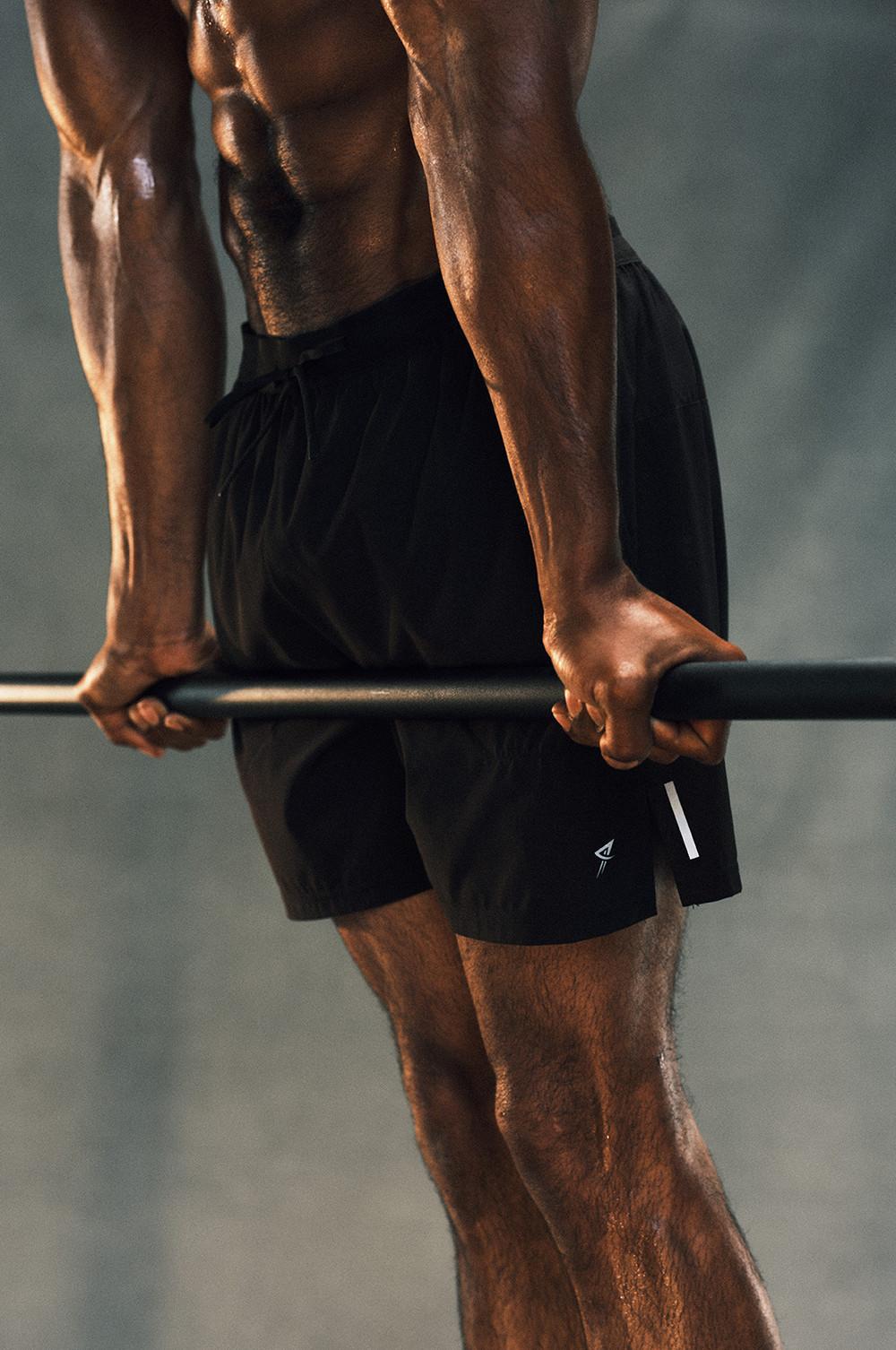 Muscular torso and arms gripping a workout bar, wearing black athletic shorts with a small logo.