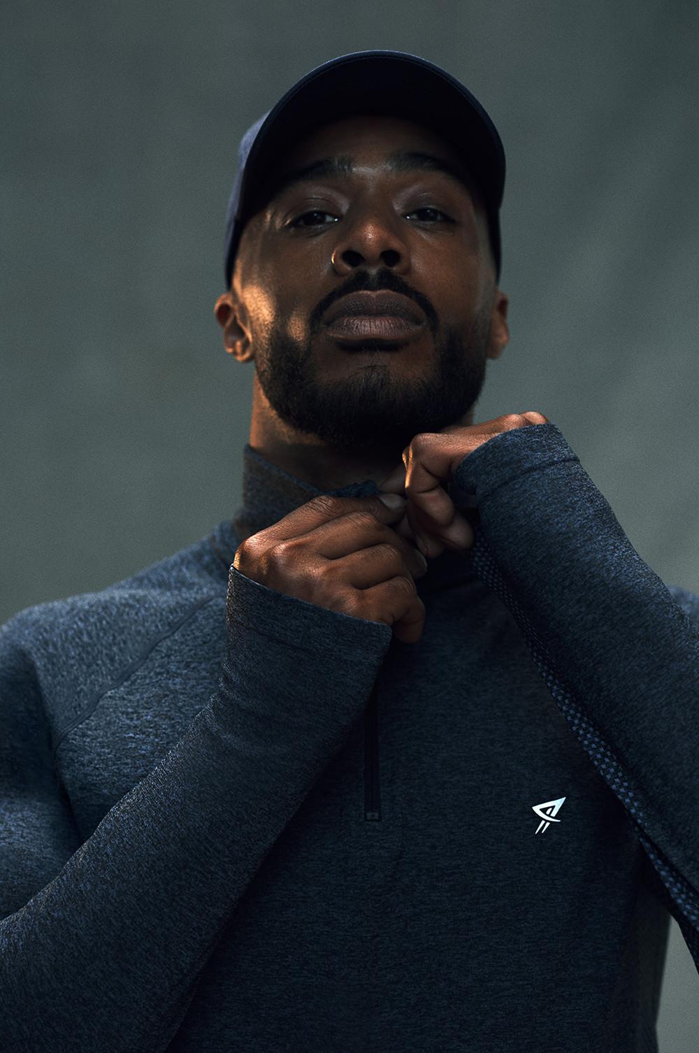 Person wearing a navy cap and grey athletic quarter-zip top with small logo, adjusting collar.