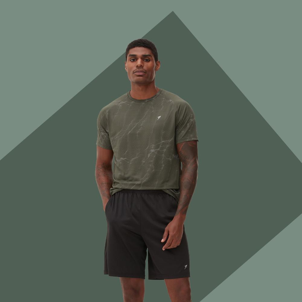 Modello che indossa una maglietta strutturata verde oliva con un piccolo logo e pantaloncini neri su sfondo geometrico verde.