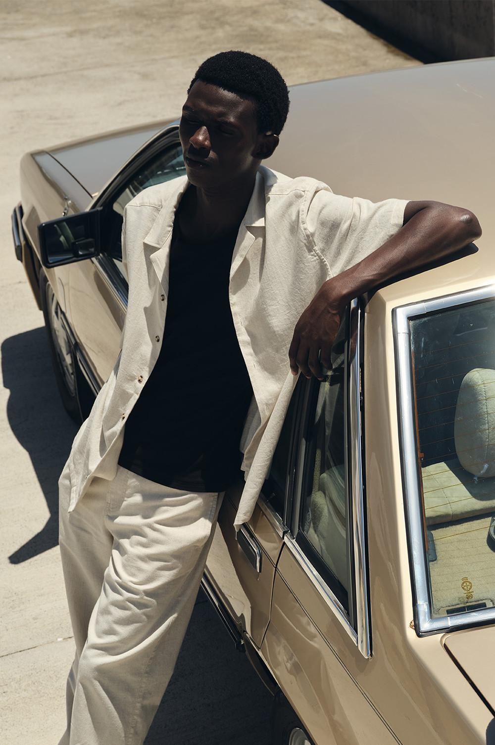Model wearing white open shirt, black t-shirt and white trousers leaning against a car