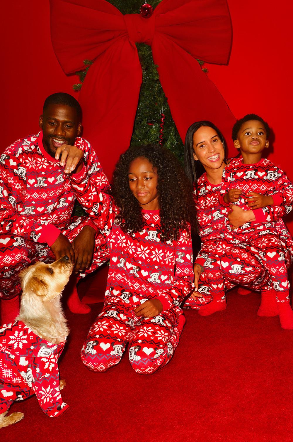 Family wearing matching Mickey Mouse Christmas pajamas with a dog against festive red backdrop with bow.