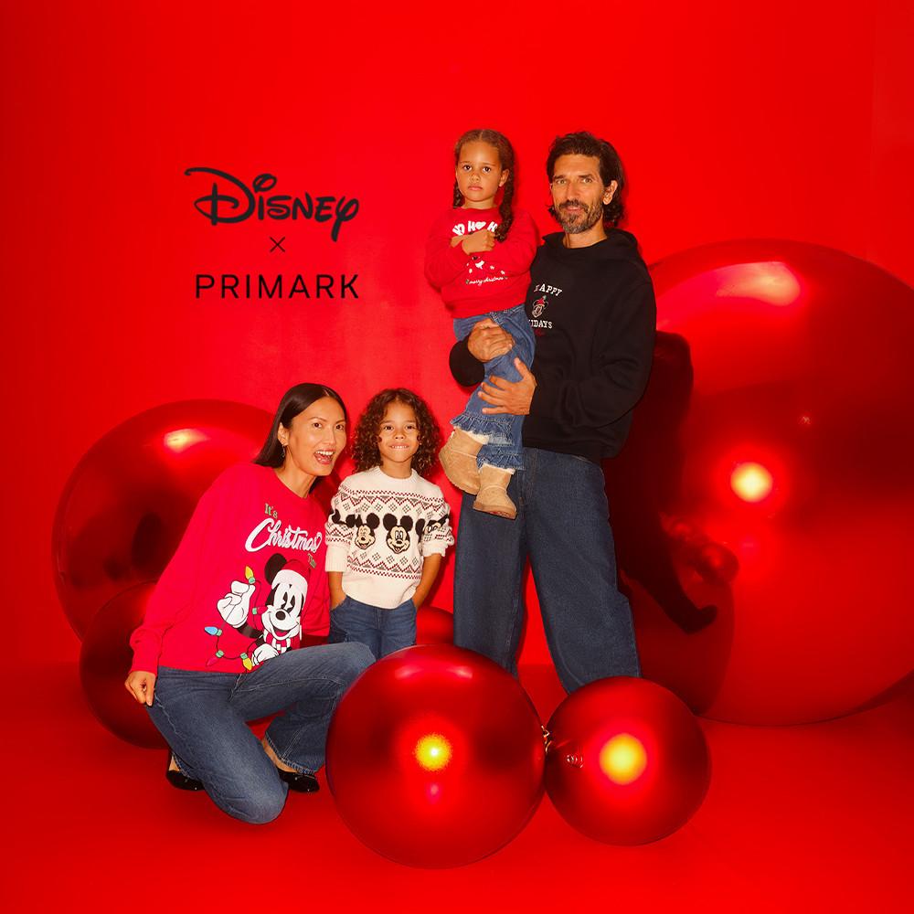 Family wearing Disney x Primark Christmas sweaters posing with large red ornaments against a red background.