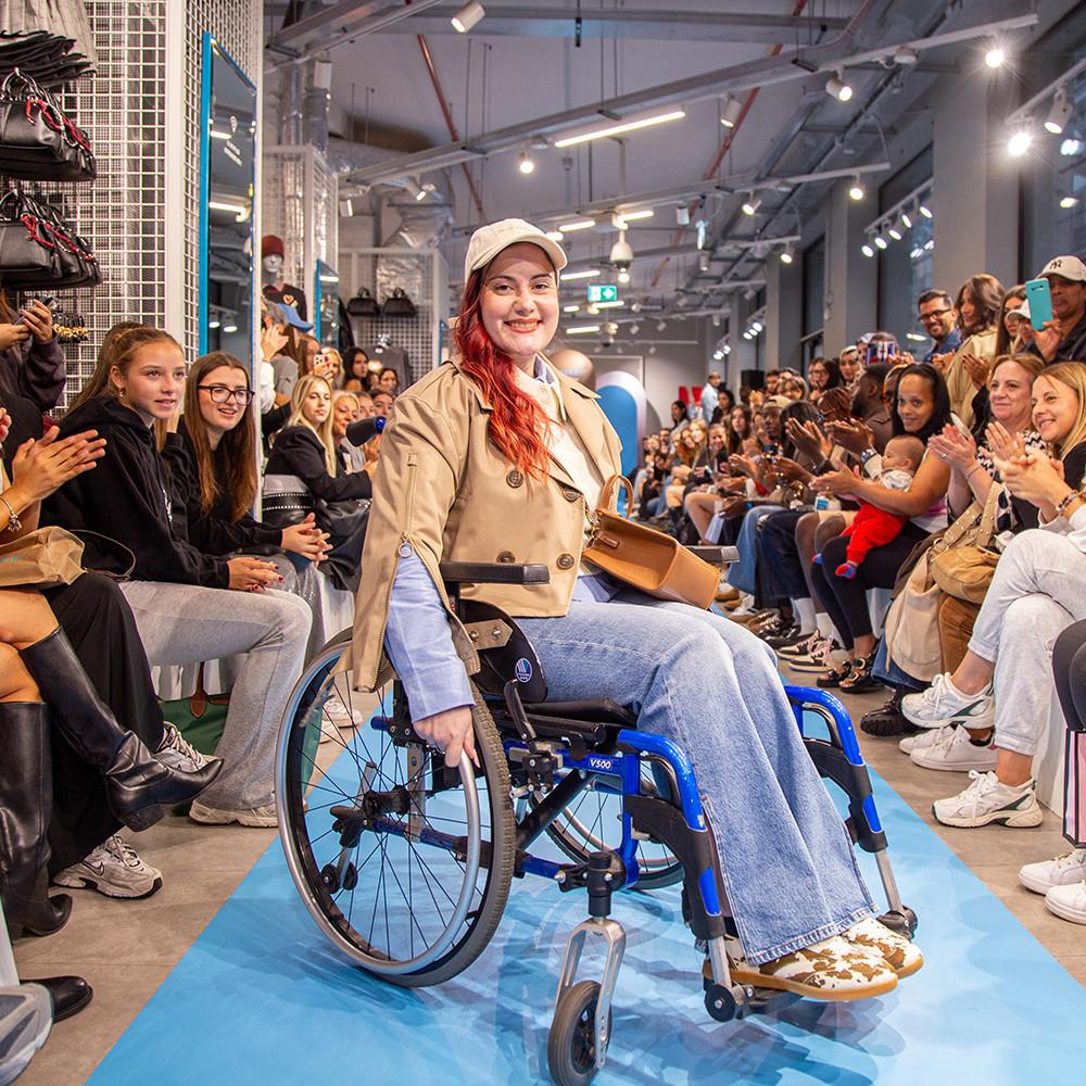 Model in wheelchair wearing beige trench coat and cap on runway with audience applauding at fashion show.