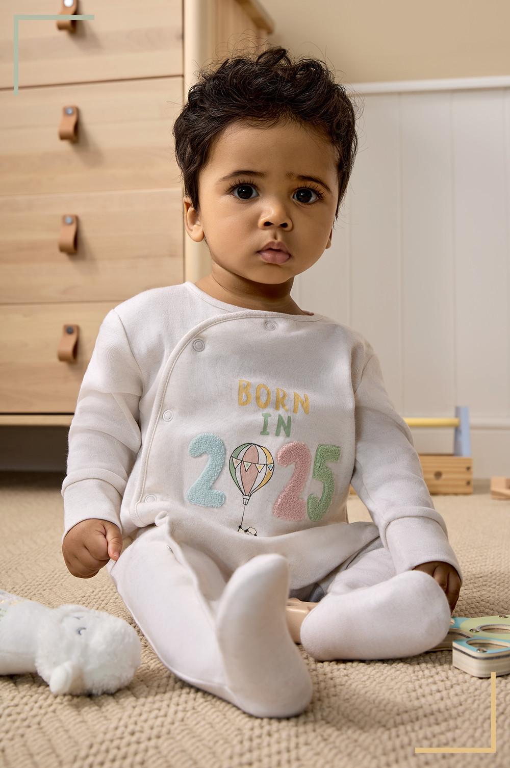 Bébé dans une grenouillère blanche avec inscription « BORN IN 2025 » et motif montgolfière, assis sur un tapis près de jouets.
