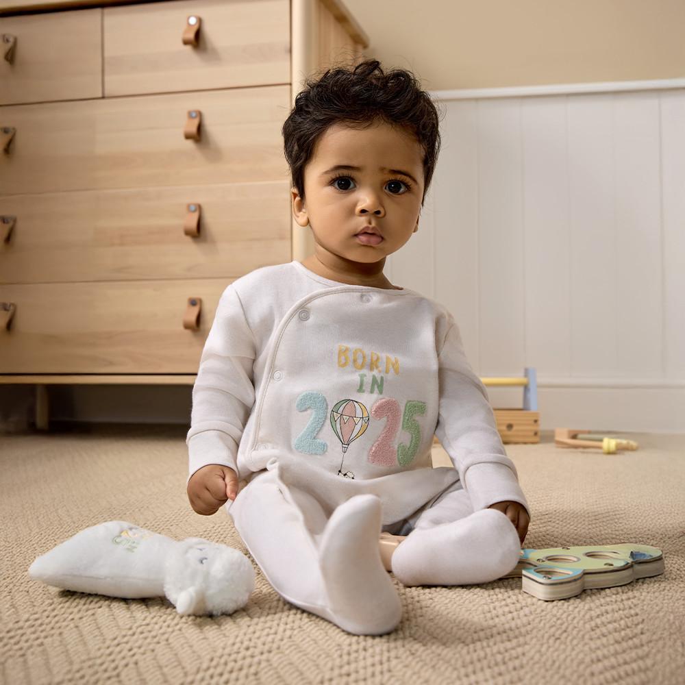 Bébé dans une grenouillère blanche avec inscription « BORN IN 2025 » et motif montgolfière, assis sur un tapis près de jouets.