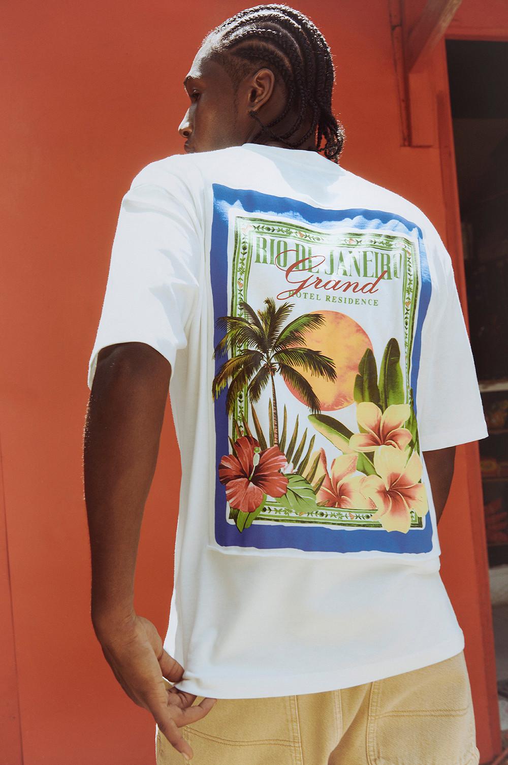 Modello che indossa una maglietta bianca a maniche corte con stampa tropicale sul retro, con fiori gialli e rosa e palme