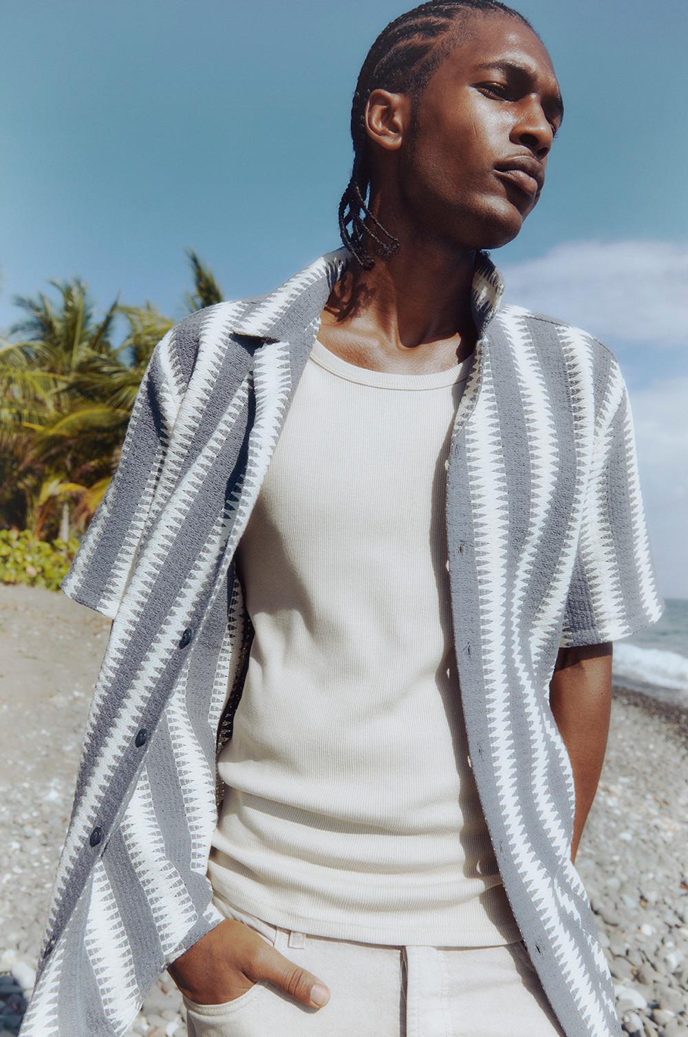 Modello che indossa una camicia all'uncinetto bianca e blu sopra una maglietta panna, insieme a un paio di pantaloncini bianchi.