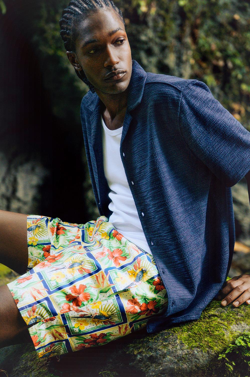 Modello che indossa una camicia blu sopra una canotta bianca, abbinato a pantaloncini con stampa a fiori tropicali