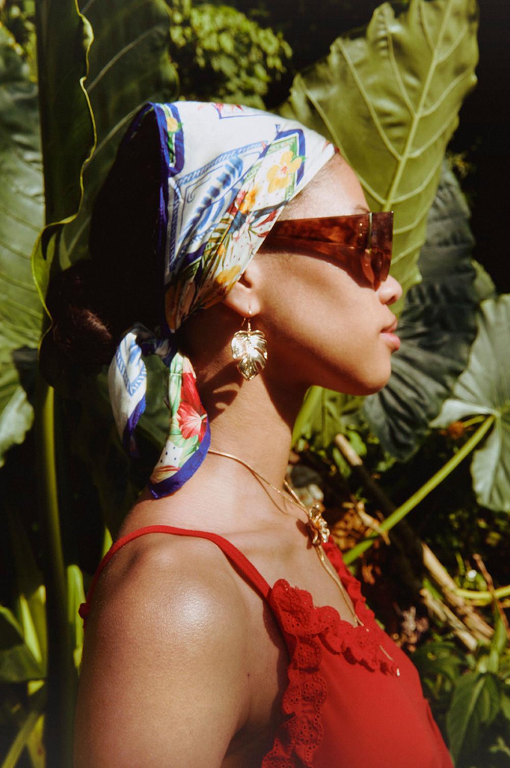 Modelo con pañuelo para la cabeza, gafas de sol marrones, pendientes dorados con forma de hoja de palmera, collar dorado y top rojo.