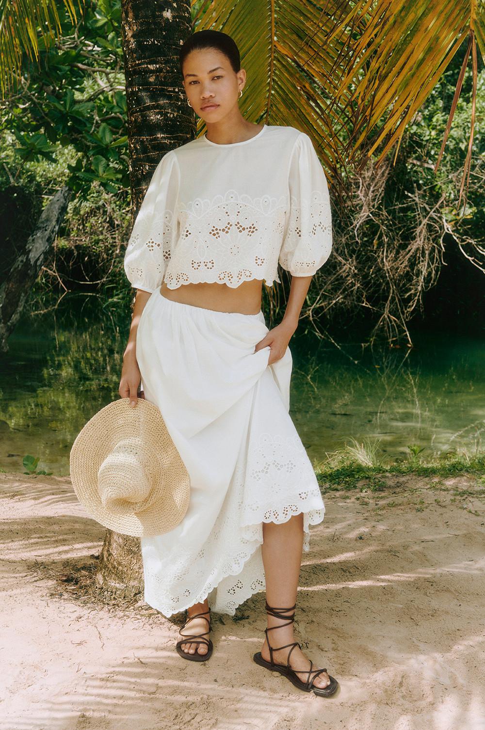 Modelo con conjunto veraniego blanco bordado con falda larga, sandalias y sombrero de paja.