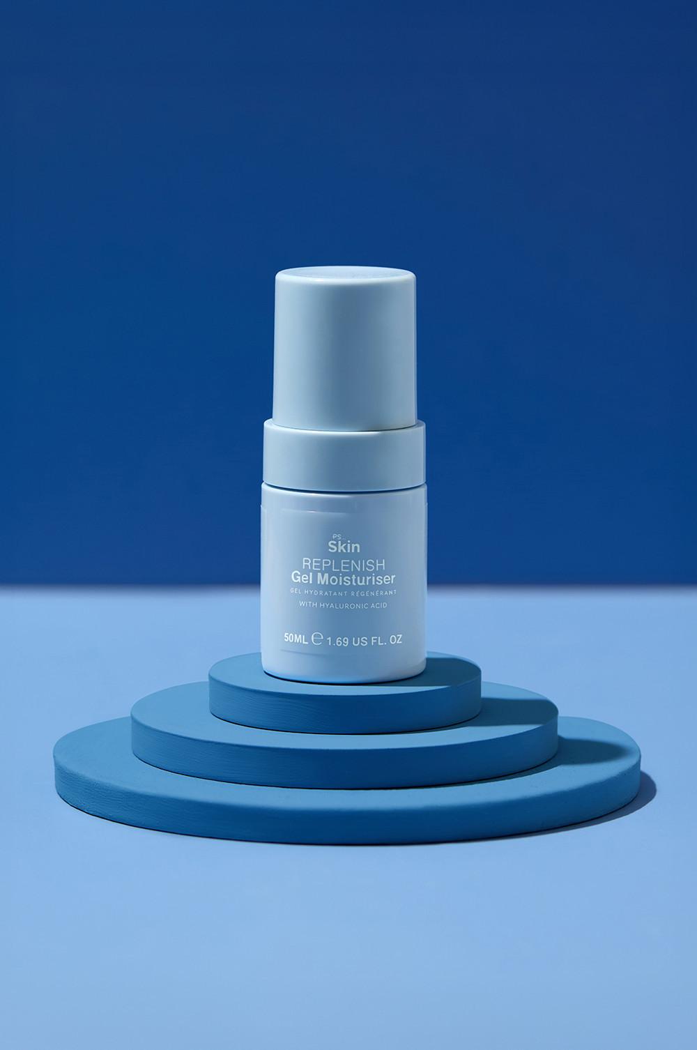 Gel hydratant Skin Replenish à l'acide hyaluronique présenté sur une plateforme bleue à étages devant un fond bleu.