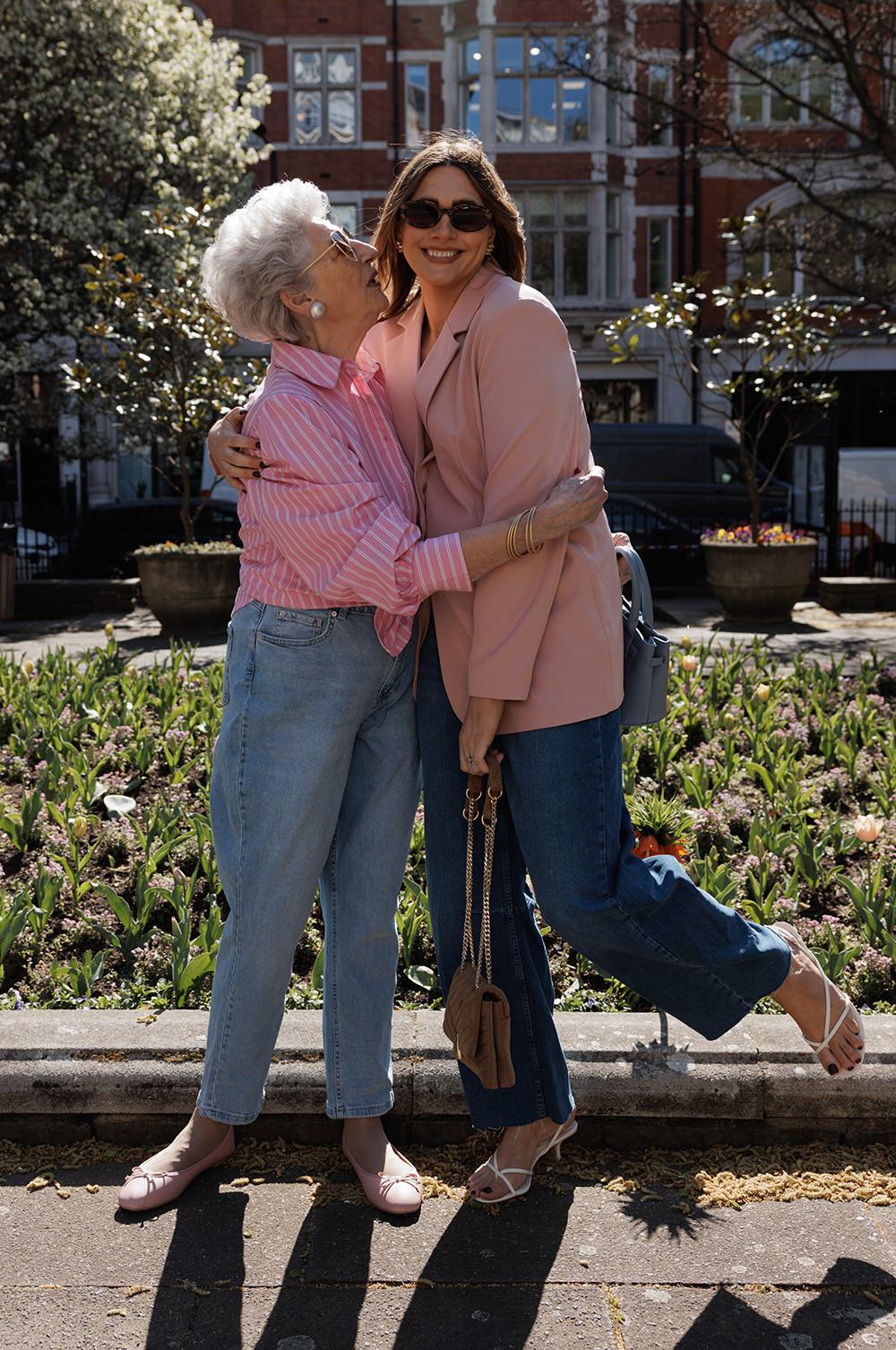 Two models, one wearing a pink blazer, jeans and heels, the other wearing a pink and white striped shirt, blue jeans and sandals