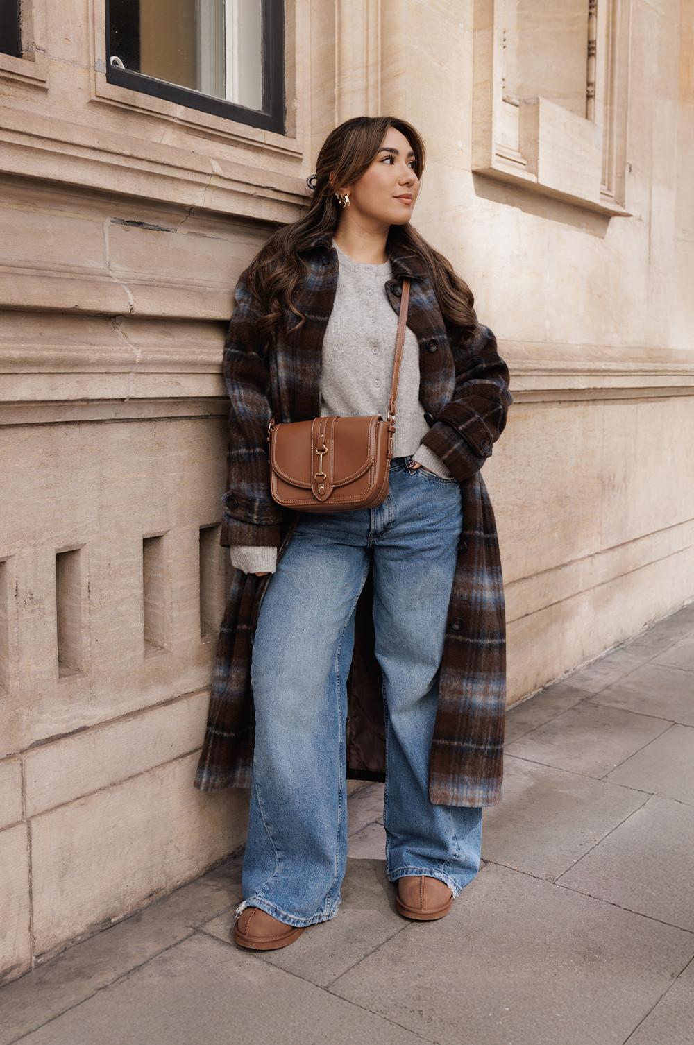Persona che indossa un cappotto con motivo scozzese sopra un maglione grigio, jeans a gamba larga e borsa a tracolla marrone davanti a un muro di pietra.