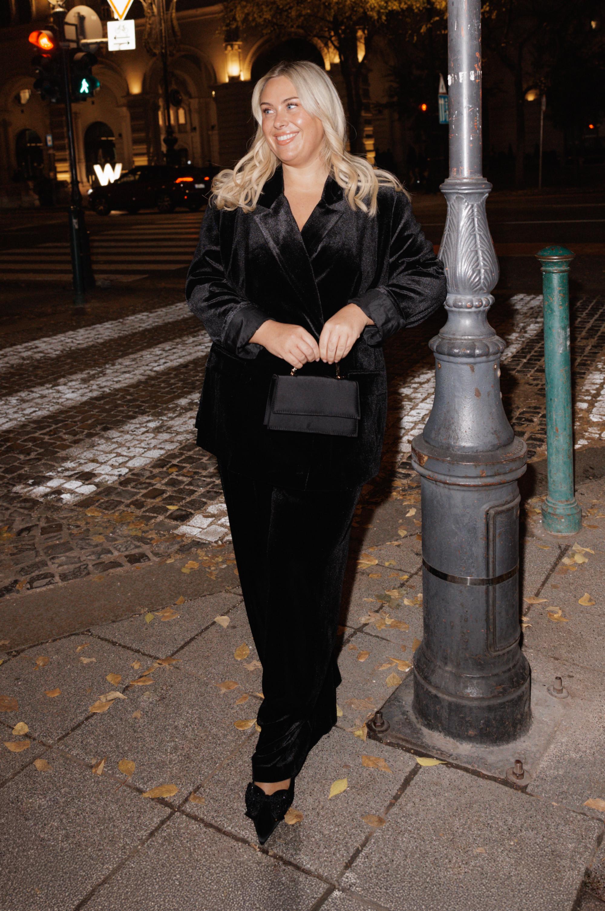 Person wearing elegant black velvet suit with matching handbag on a city street at night.