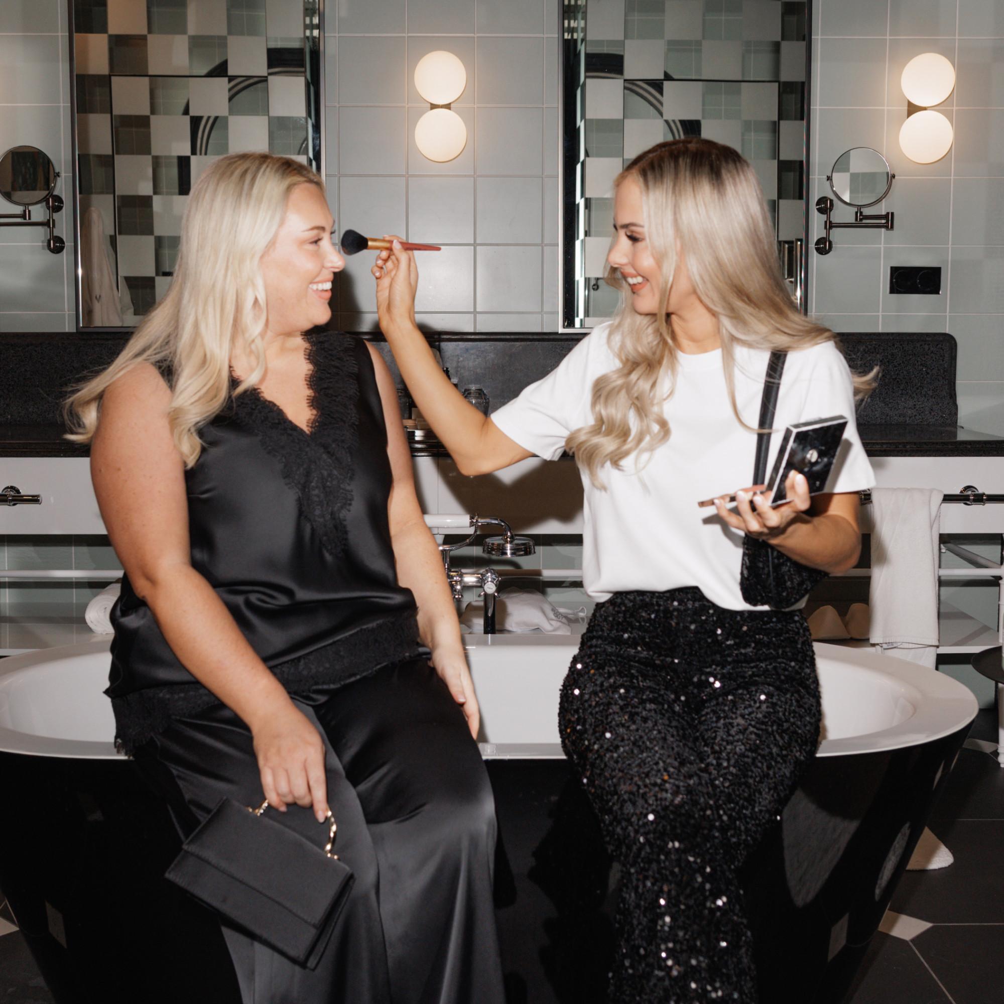 Two women in stylish outfits applying makeup in a modern bathroom, one holding a makeup brush and palette.
