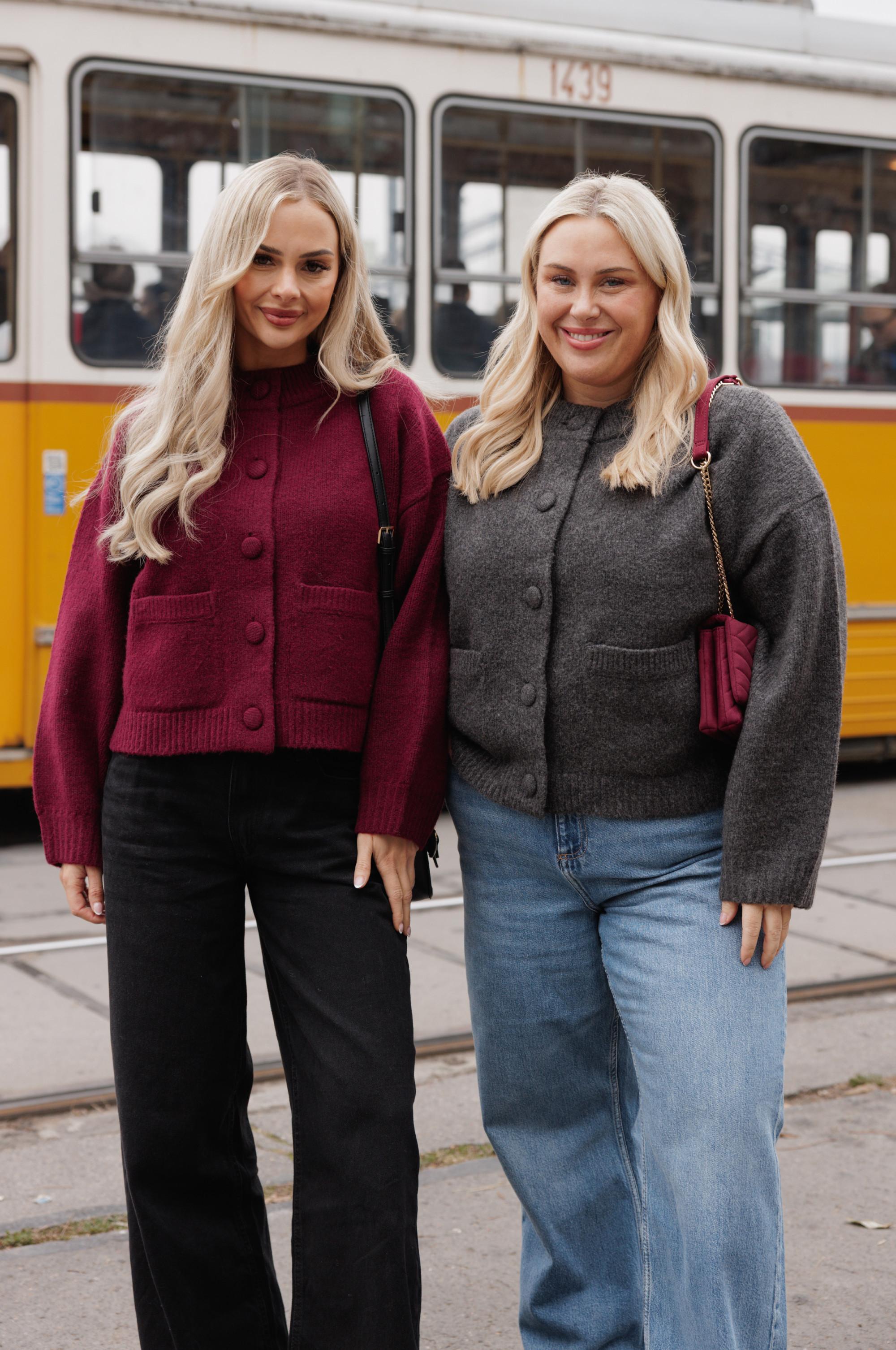 Two women wearing stylish button-up cardigans - burgundy and grey - standing in front of a yellow tram.