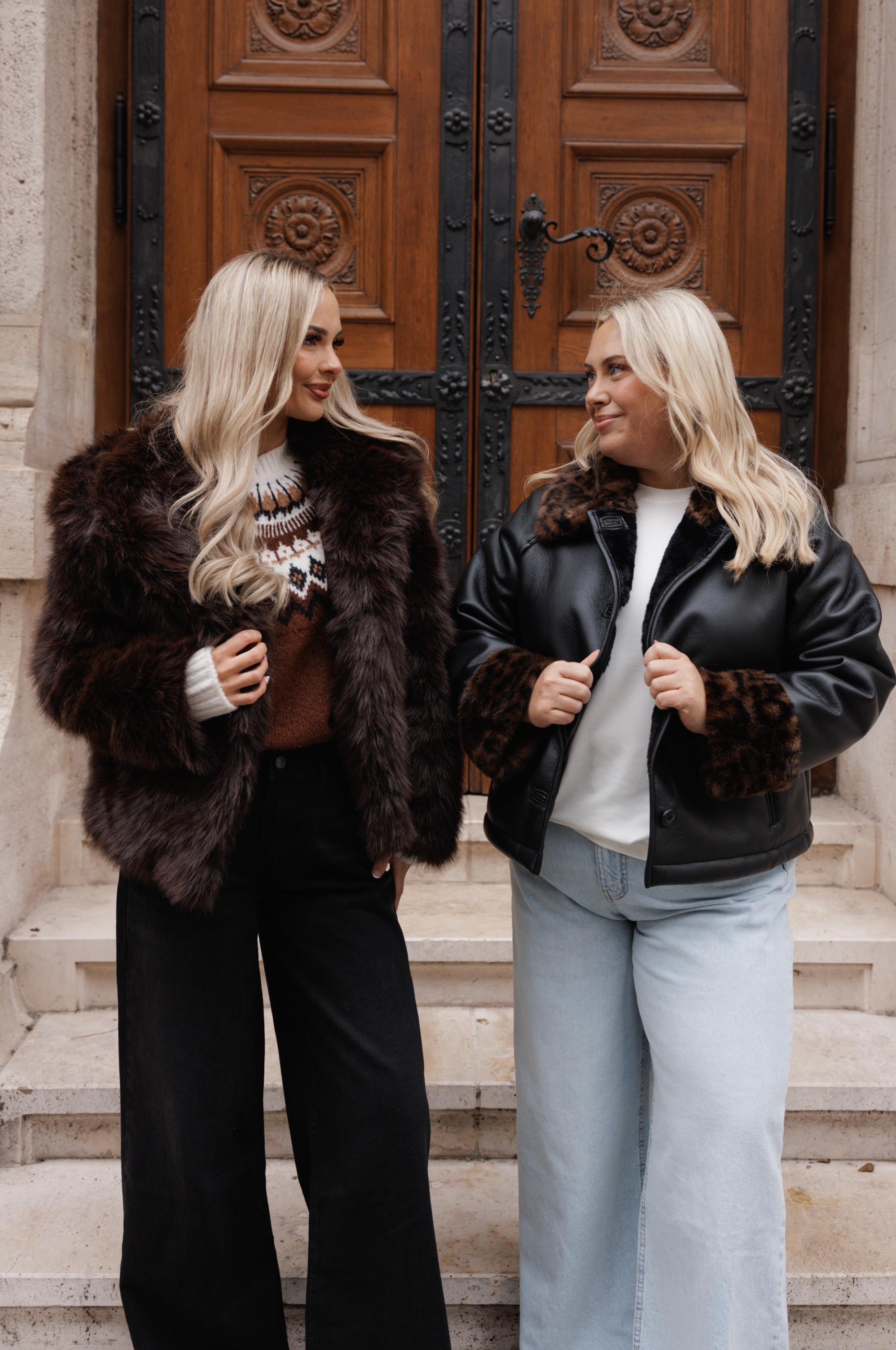 Two women wearing winter outerwear - brown fur coat and black leather jacket with fur trim - standing in front of ornate wooden doors.