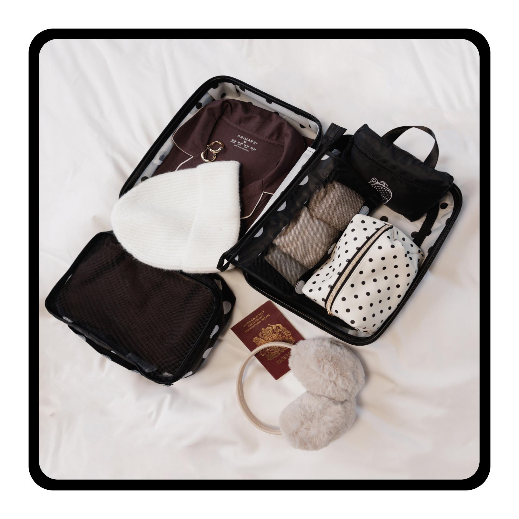 Open suitcase with neatly packed clothing, toiletry bags, a white beanie, fluffy earmuffs and British passport on white bedding.