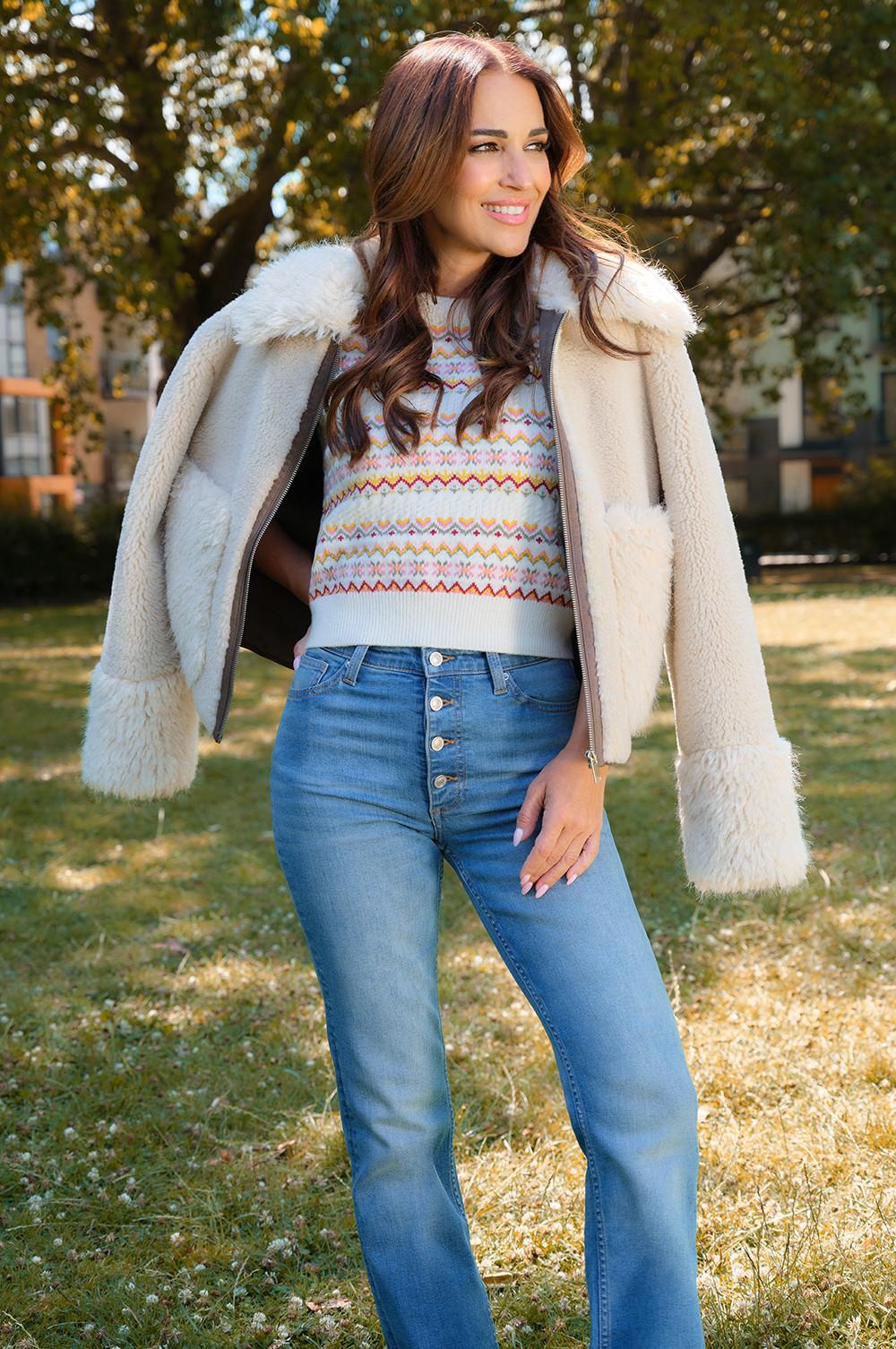 Person wearing cream sherpa jacket over patterned knit jumper and blue jeans in an autumn outdoor setting.