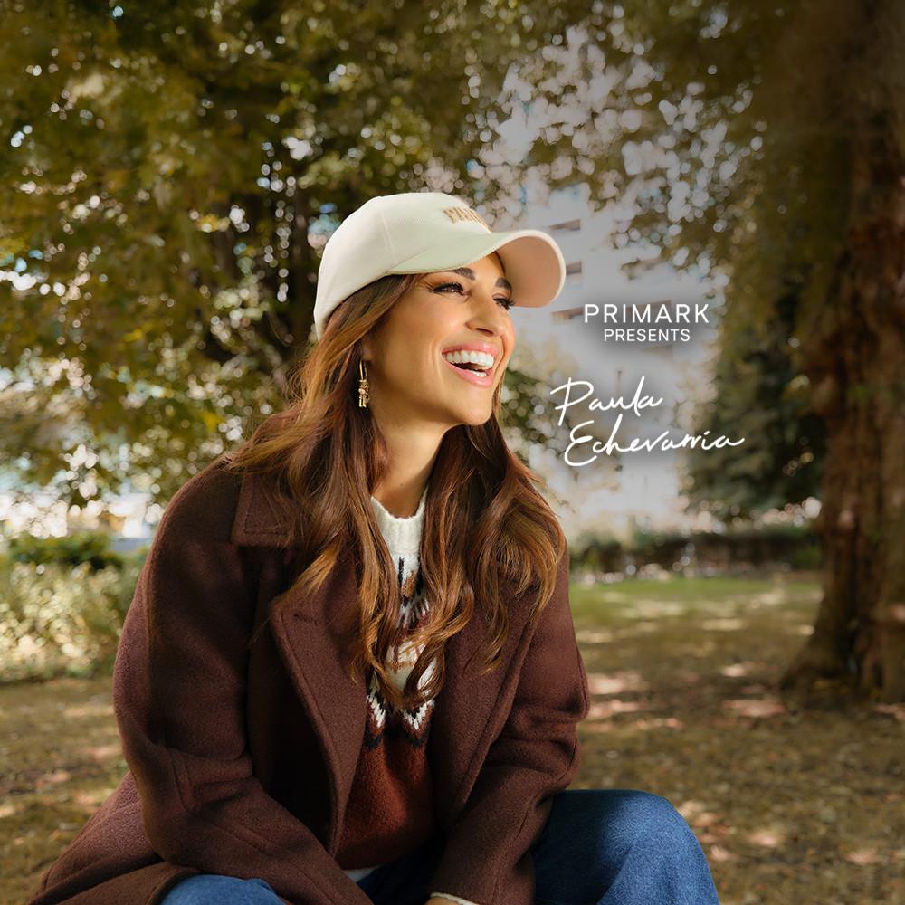 Person wearing a cream cap and brown coat in an autumn park setting, Primark fashion advertisement.
