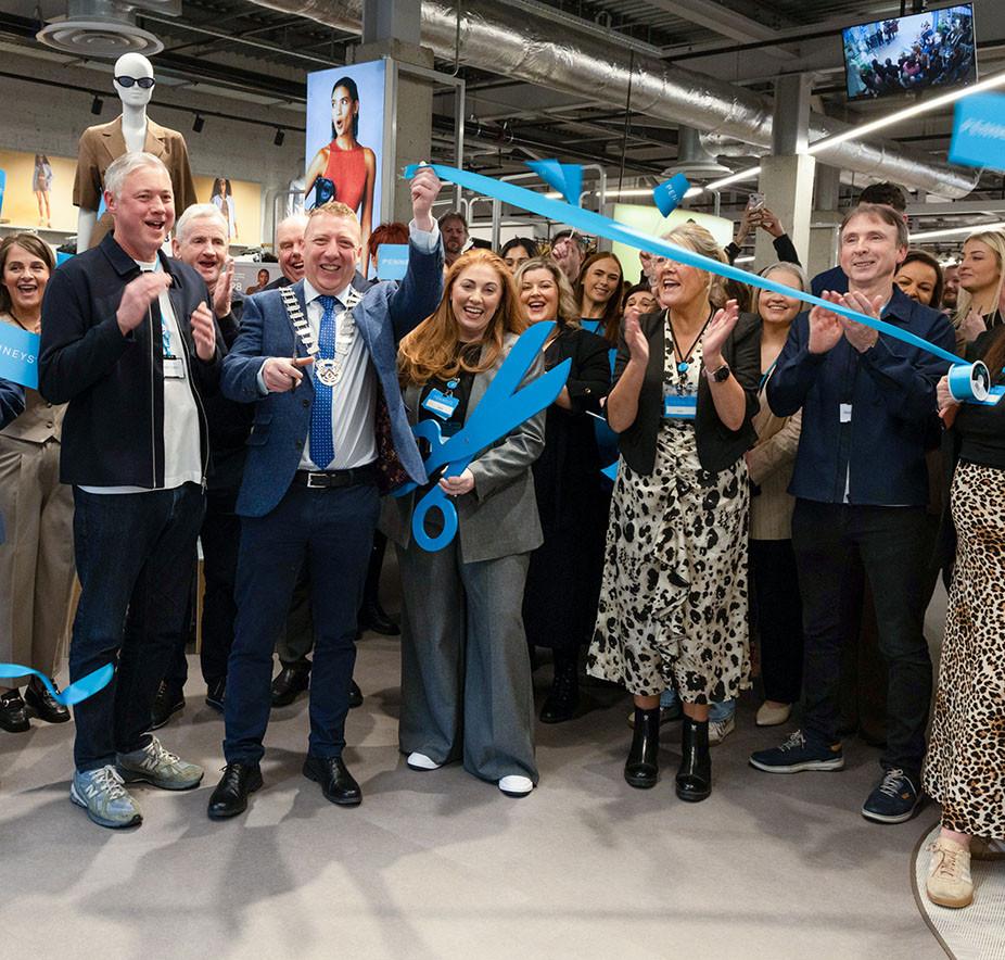 A group of people cheer as a man with a chain of office cuts a blue ribbon with oversized scissors.