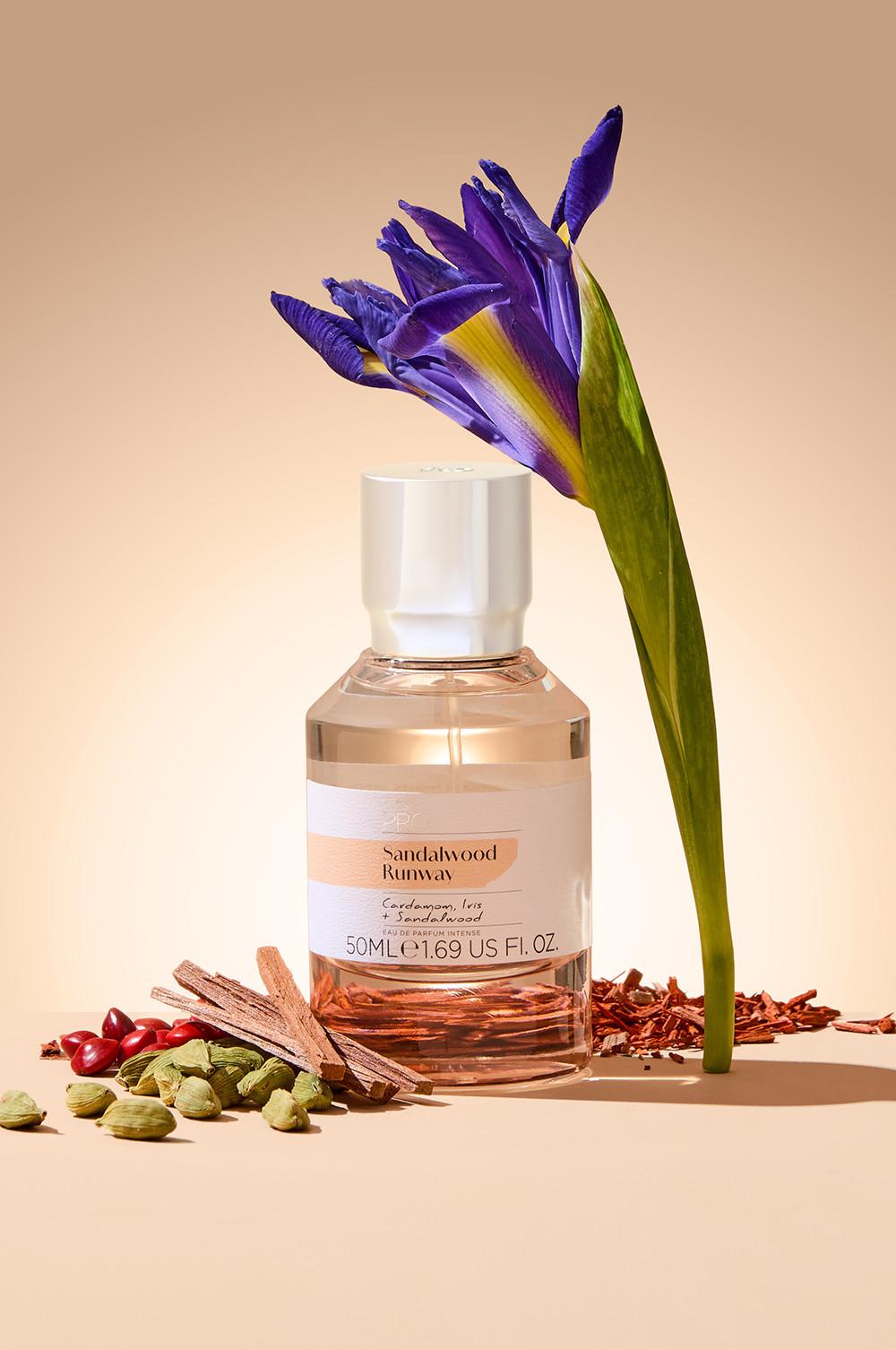 Flacon de parfum Sandalwood Runway présenté entouré d'un iris violet, de capsules de cardamome et d'épices disposés avec créativité devant un fond neutre. Format 50 ml.