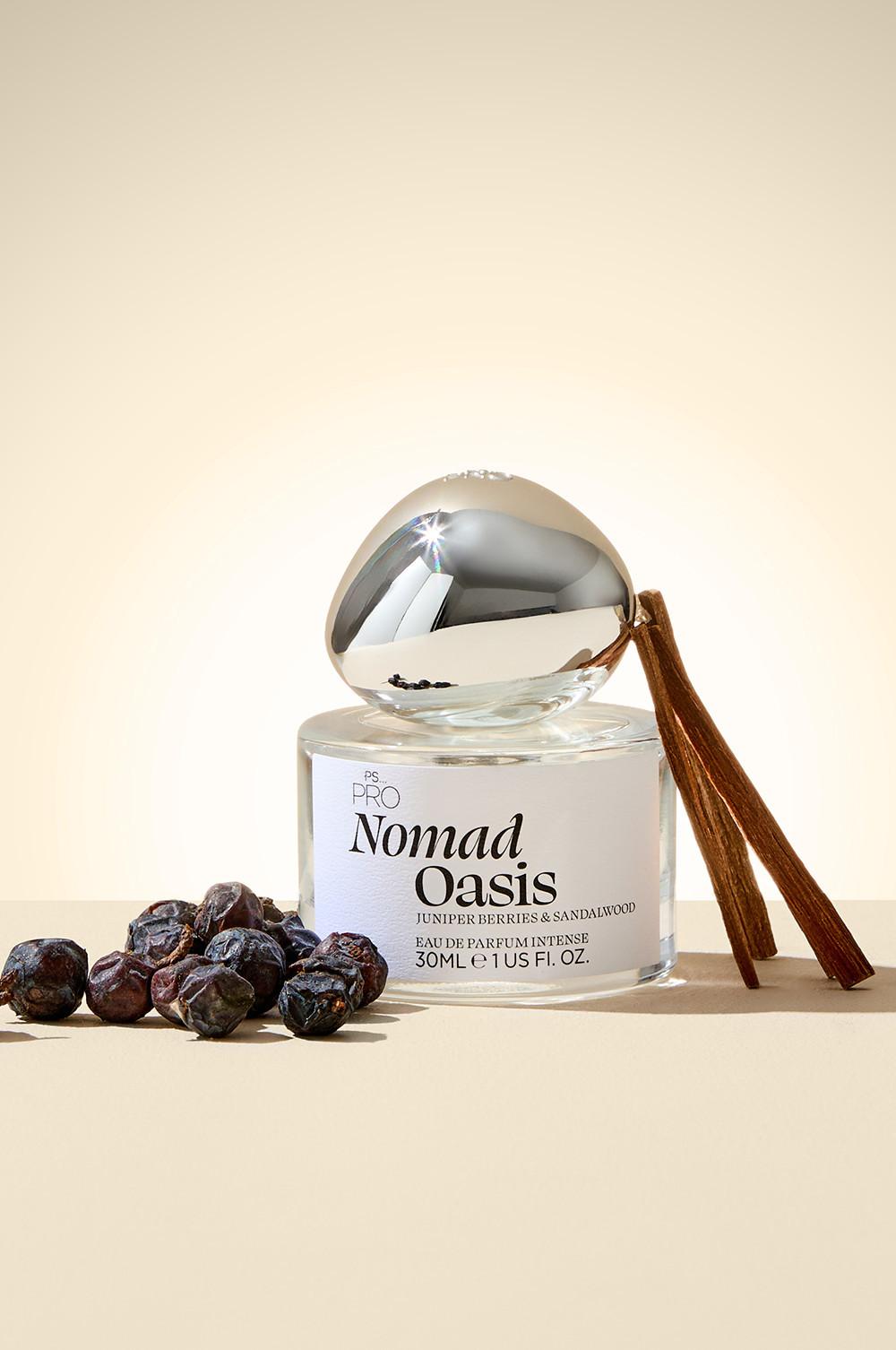 Flacon de parfum Nomad Oasis de PS... PRO doté d'un capuchon argenté, aux notes de baies de genévrier et de bois de santal, présenté devant des bâtons de cannelle et des baies séchées.