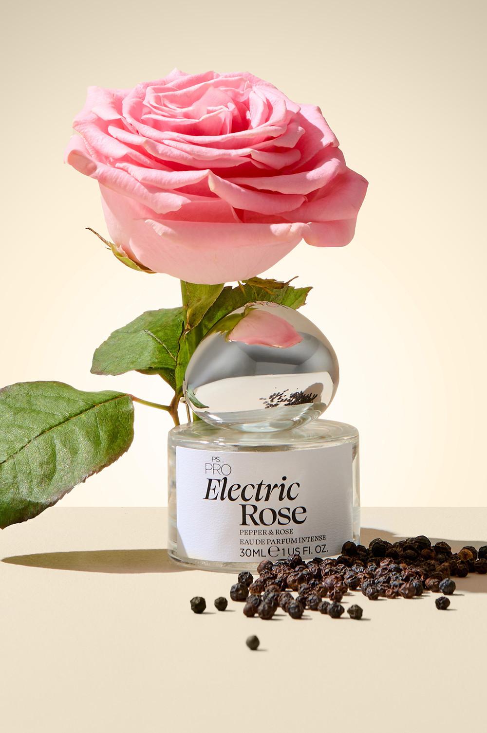 Flacon de parfum Electric Rose de PS... PRO présenté avec une rose rose sur le dessus et des grains de poivre parsemés tout autour.
