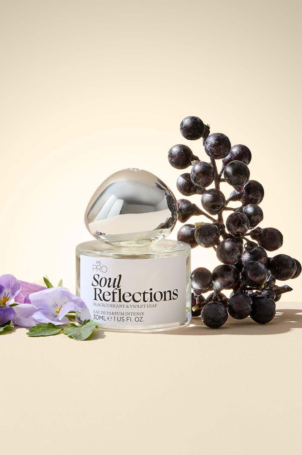 Flacon de parfum Soul Reflections de PS... PRO aux notes de cassis et de violette présenté devant des baies fraîches et des fleurs violettes.