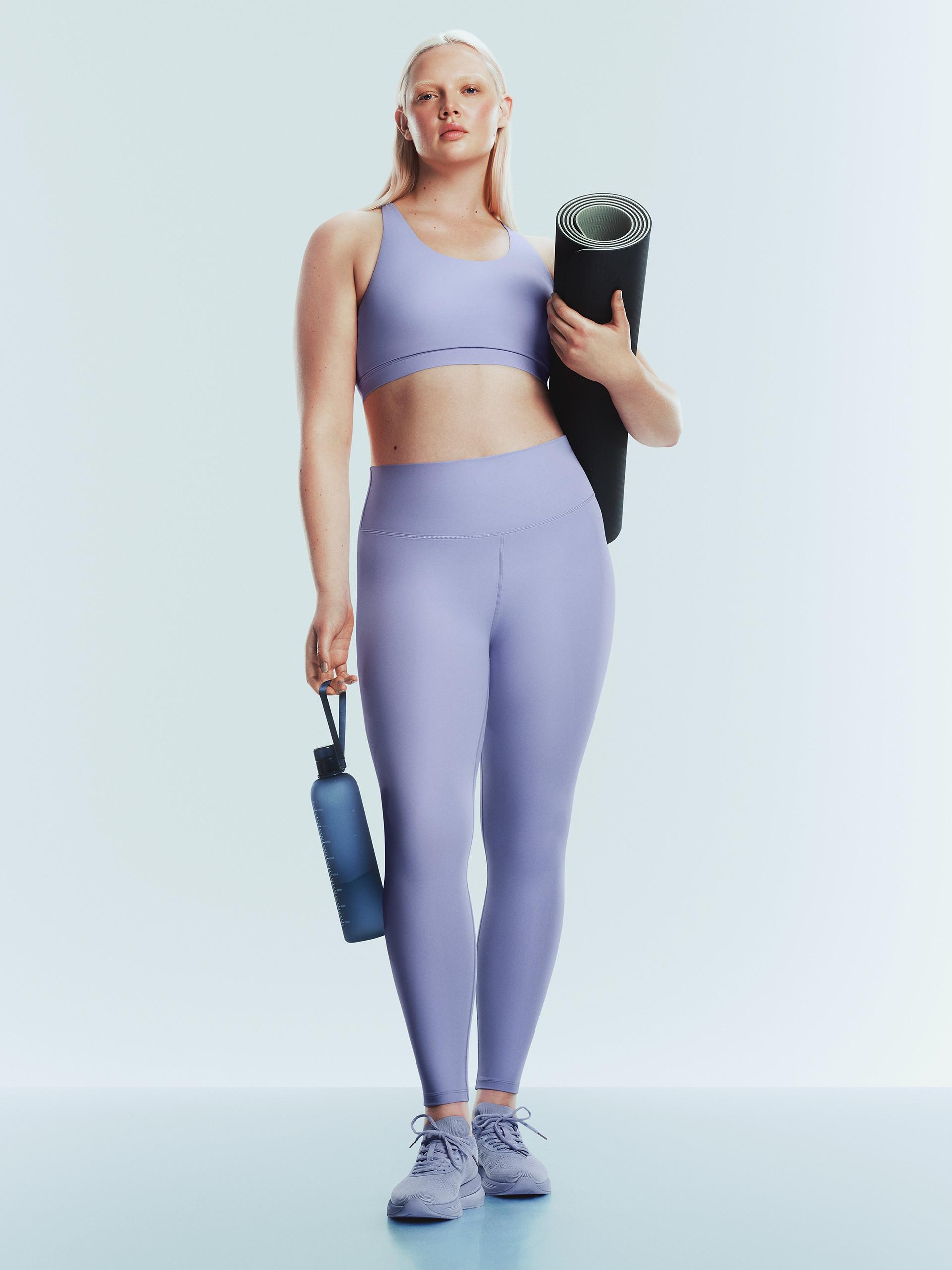 Person in pink fitted activewear holding a yoga mat and water bottle, balancing on one leg against white background.