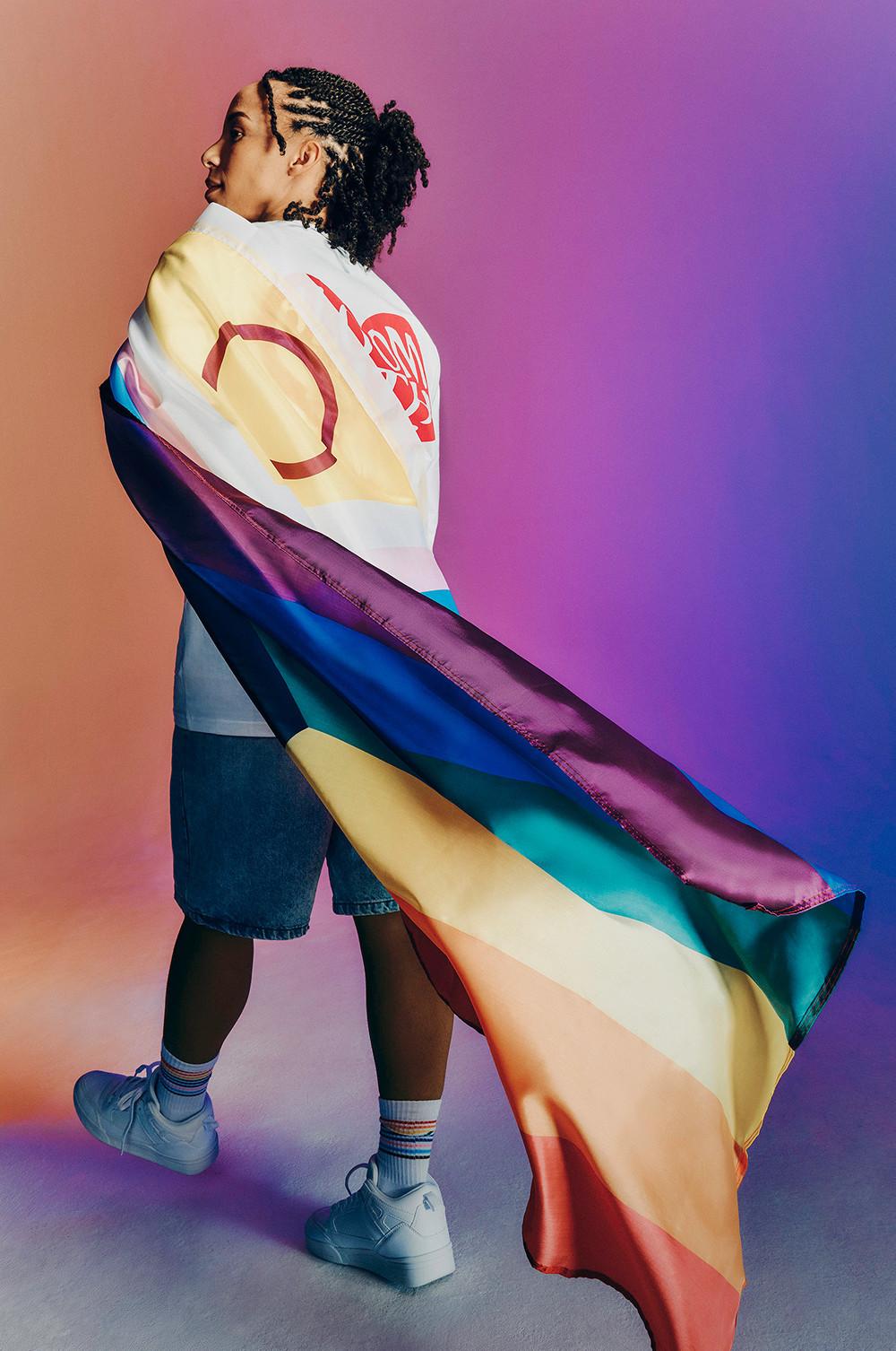 Modella che indossa una maglietta bianca “Bloom with Pride”, con la bandiera del Pride sulle spalle.