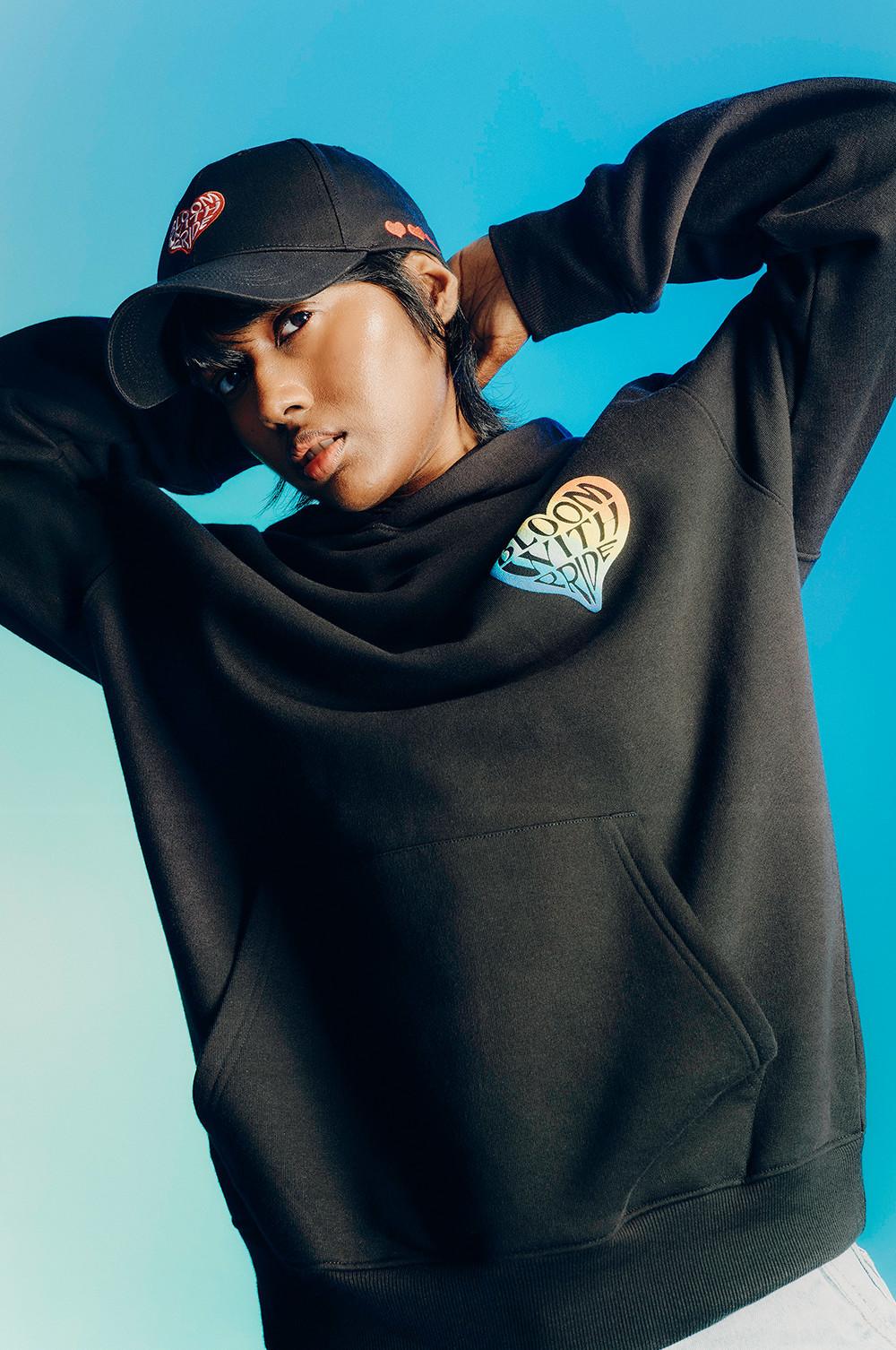 Modella che indossa una felpa nera con cappuccio con il logo “bloom with pride” sul davanti e un cappellino nero abbinato.