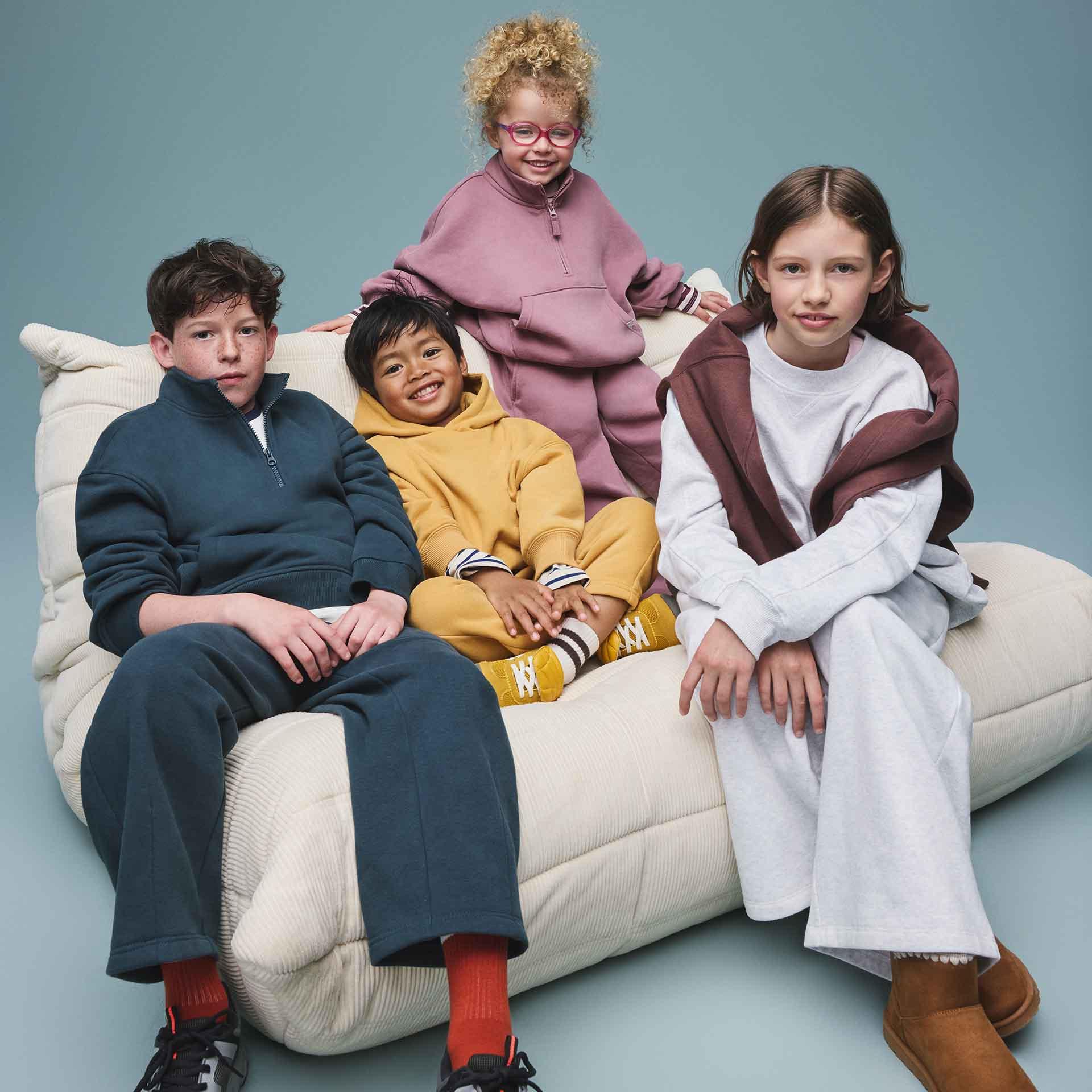 Four children sitting on a cream-colored sofa wearing colourful sweatshirts, hoodies, and joggers.