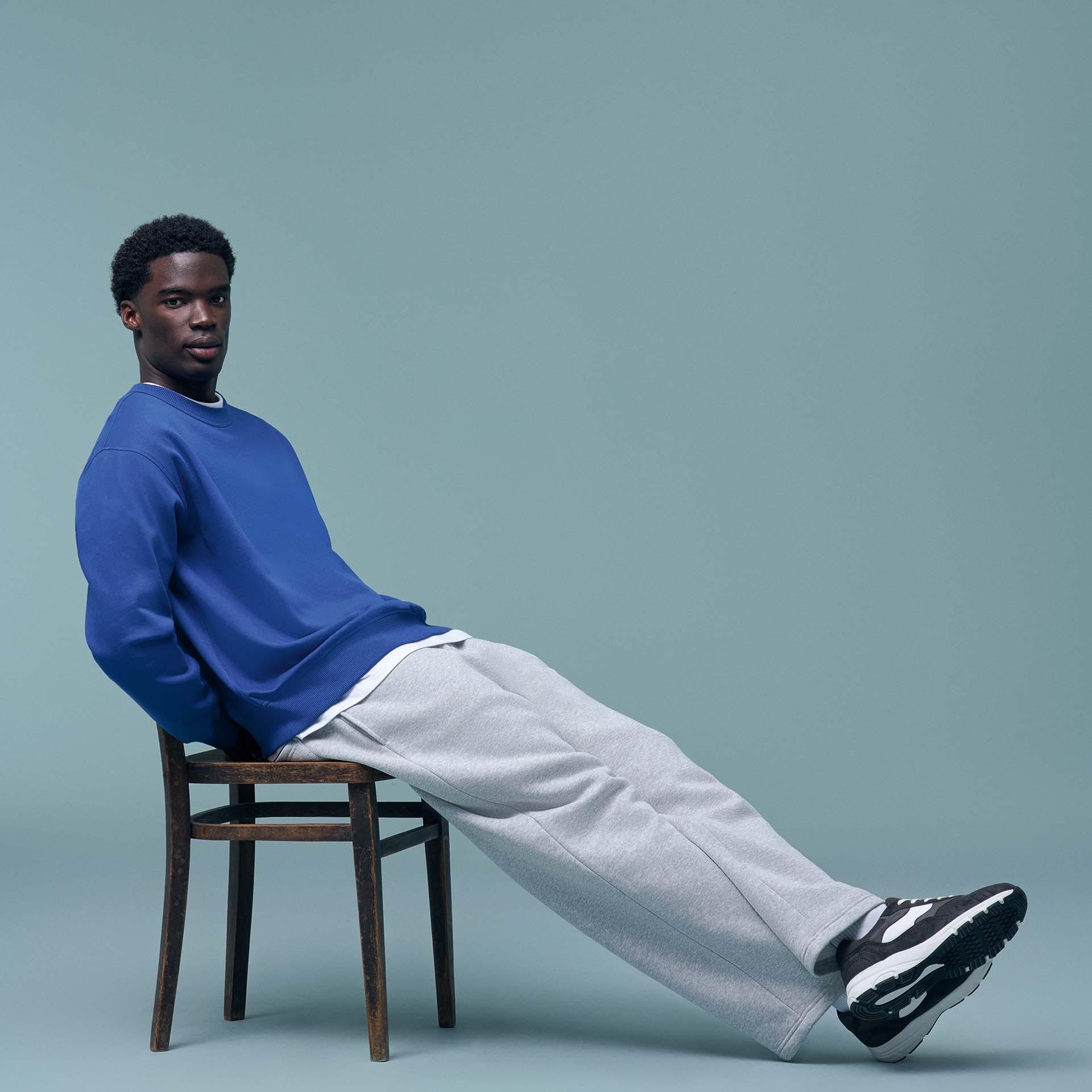 A young man wearing a blue crewneck sweatshirt and grey joggers sits on a wooden chair.