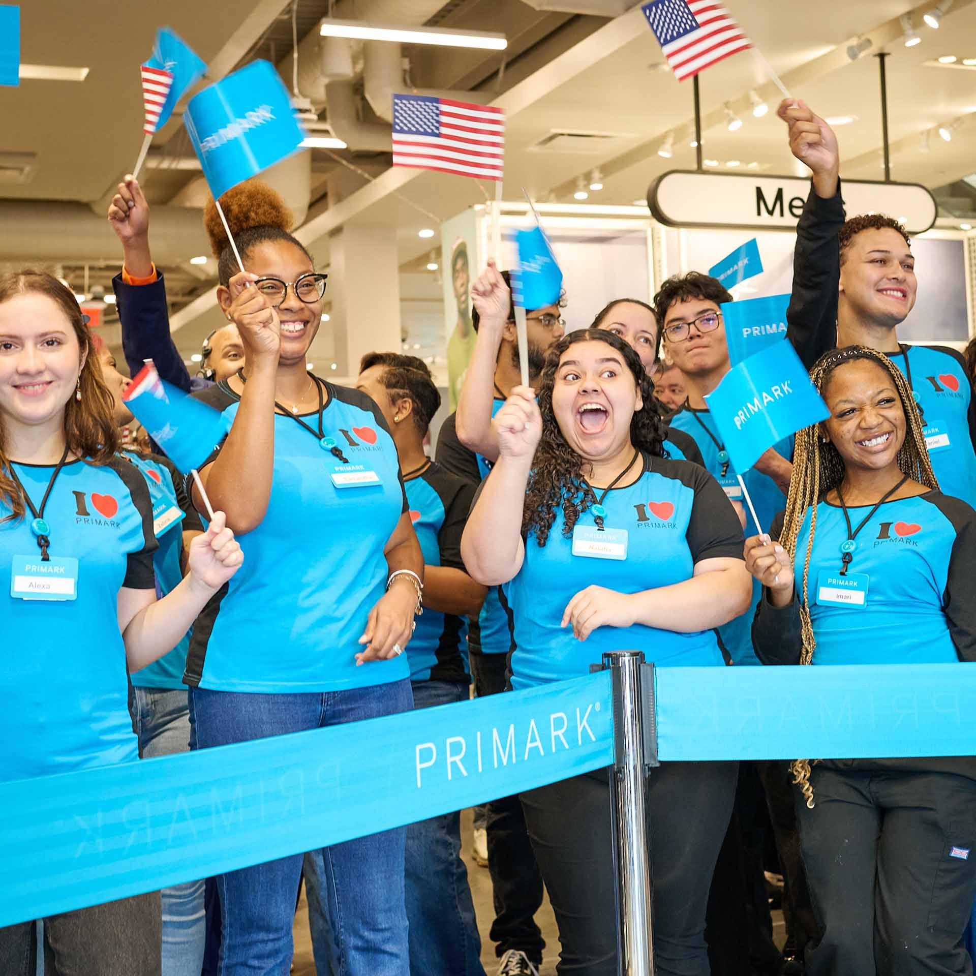 Smiling Primark employees hold American and Primark flags behind a blue store barrier.