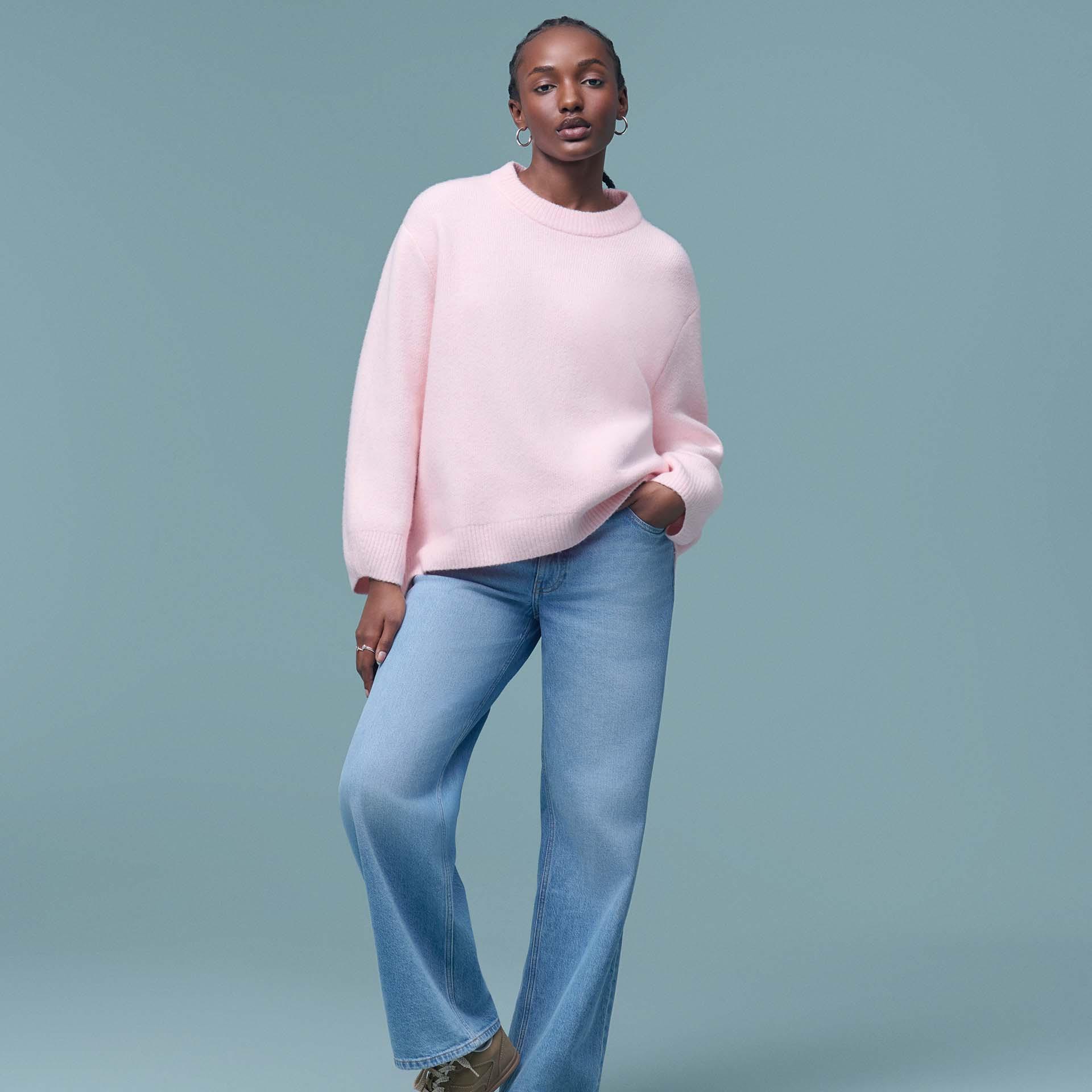 A woman with braided hair wears a light pink crewneck sweater and light blue wide-leg denim jeans.