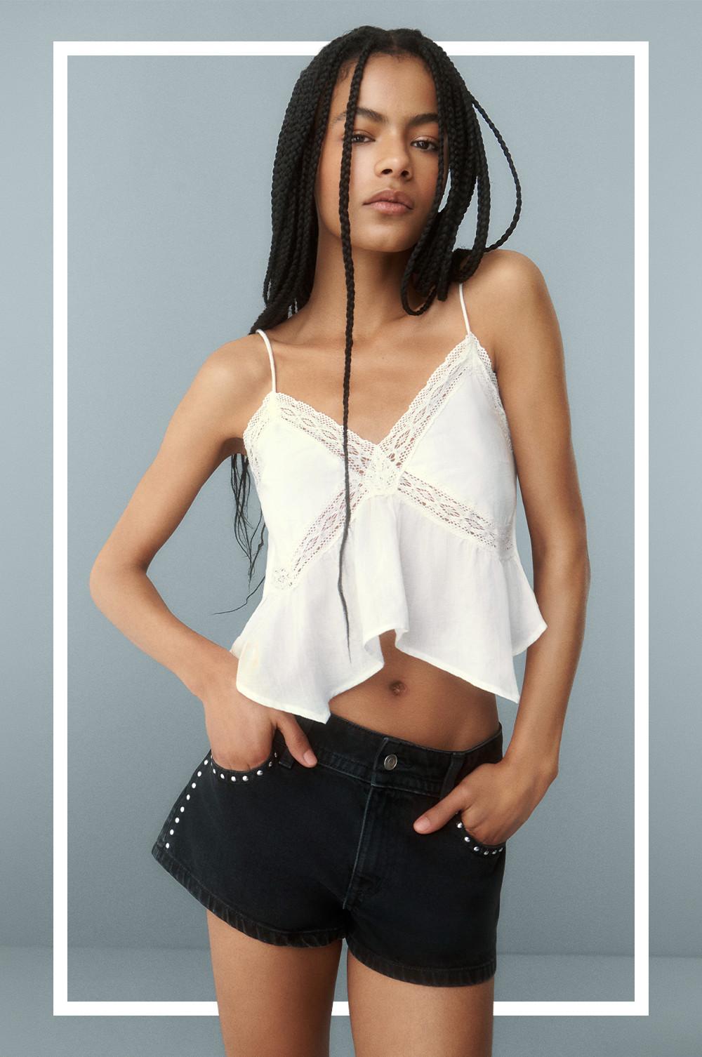 Model wearing white lace-trimmed cami top with asymmetric hem and black studded denim shorts.